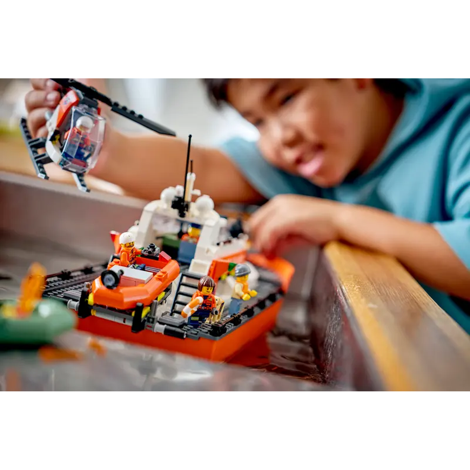 A child plays with a Lego boat and helicopter. The boat is in water, with orange and white colors. The helicopter is held by the child.