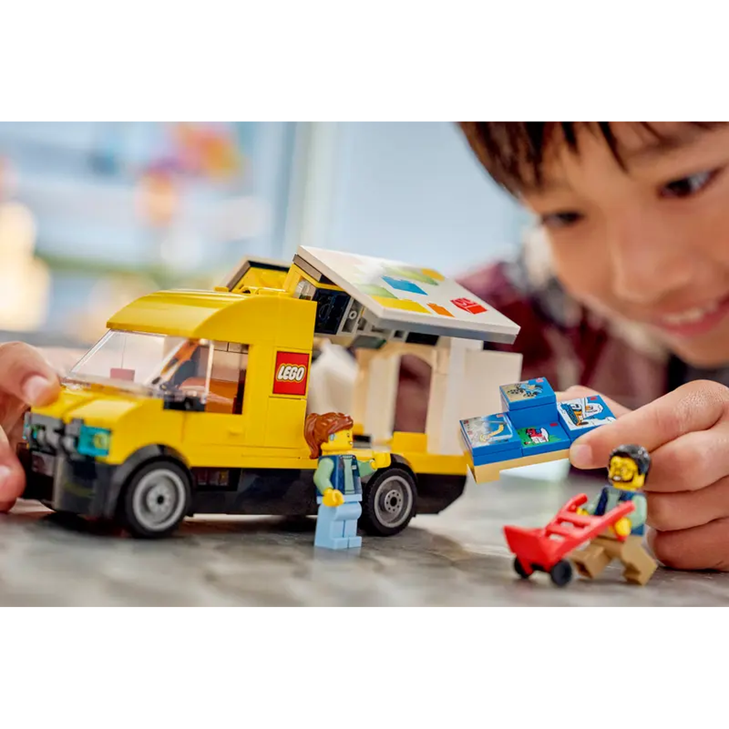 A child plays with a Lego food truck. The yellow truck has its roof open. Two Lego figures stand near the truck and another pulls a red cart.