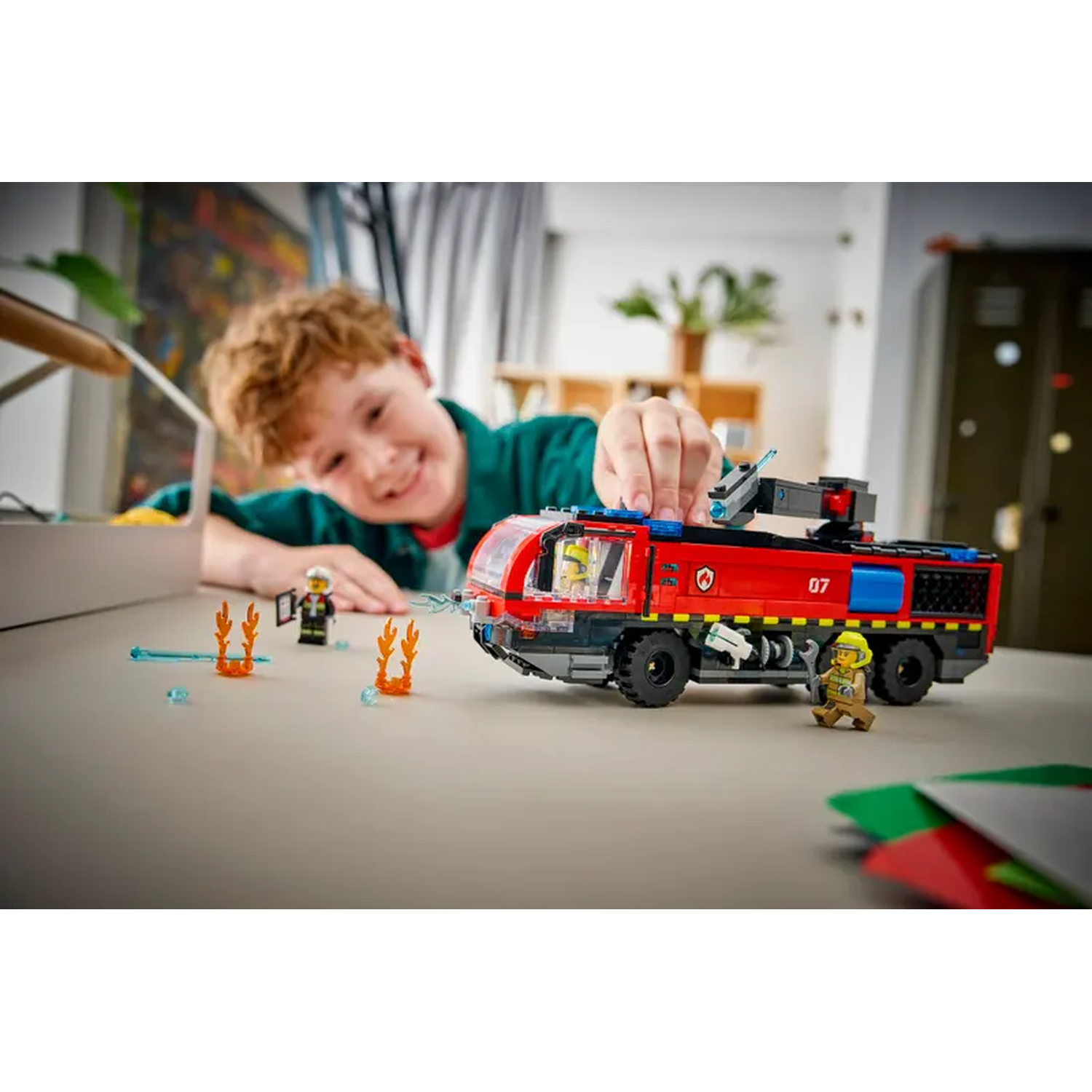 A boy smiles, playing with a LEGO fire truck. Firefighters and flames are on the table. The truck is red with black details.