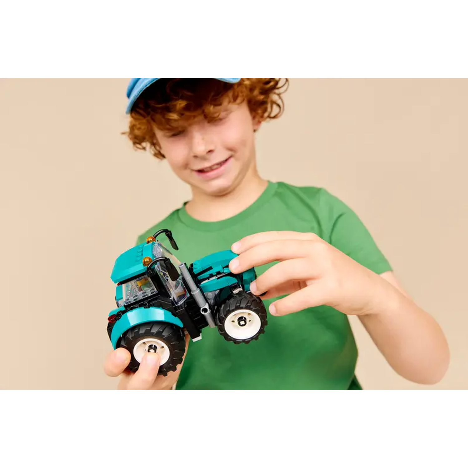 A boy holds a teal toy tractor. He wears a blue cap and a green shirt. Background is a light beige.