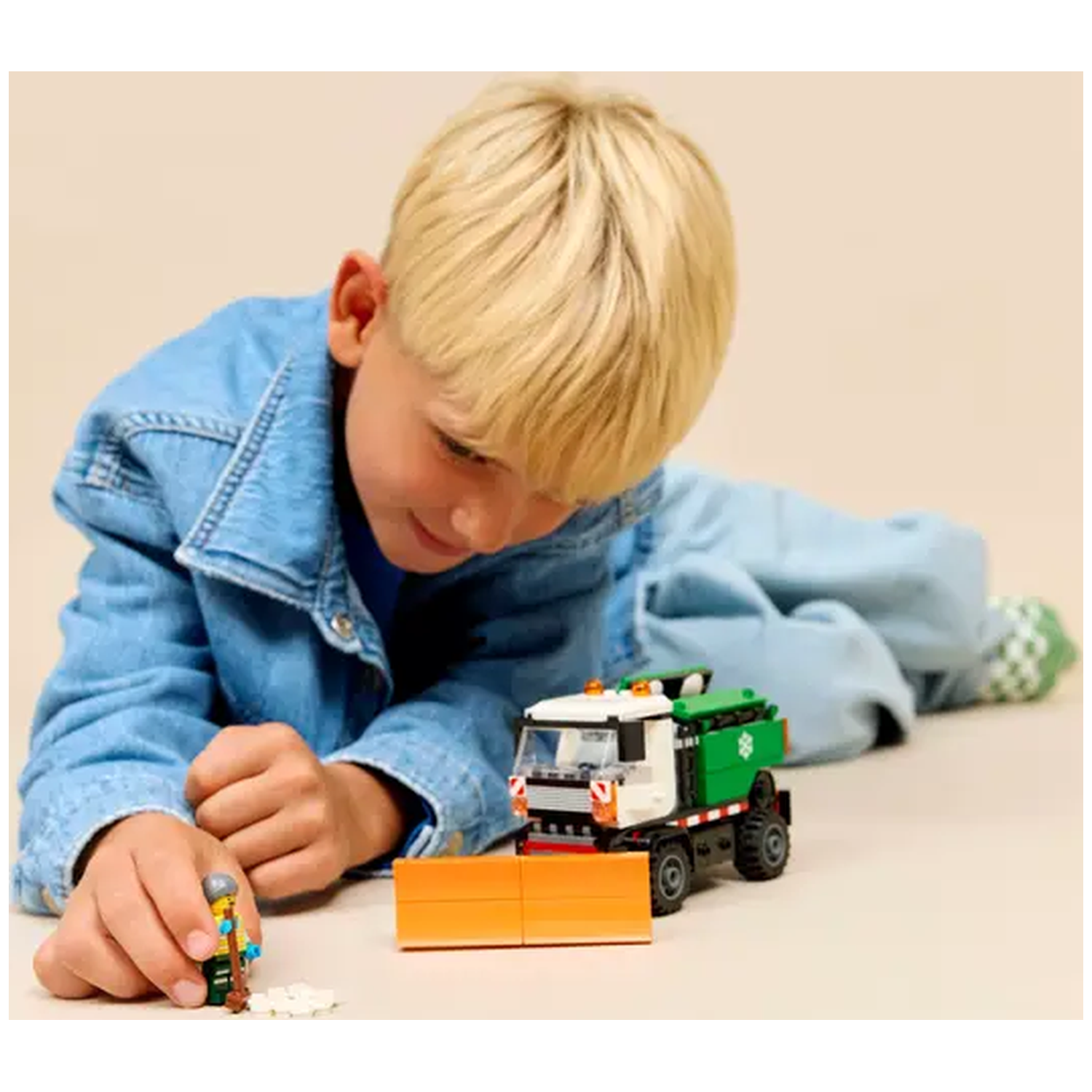 A boy in denim plays with a Lego snowplow.  He holds a Lego figure with a shovel.  A snowplow and snow are in the foreground.