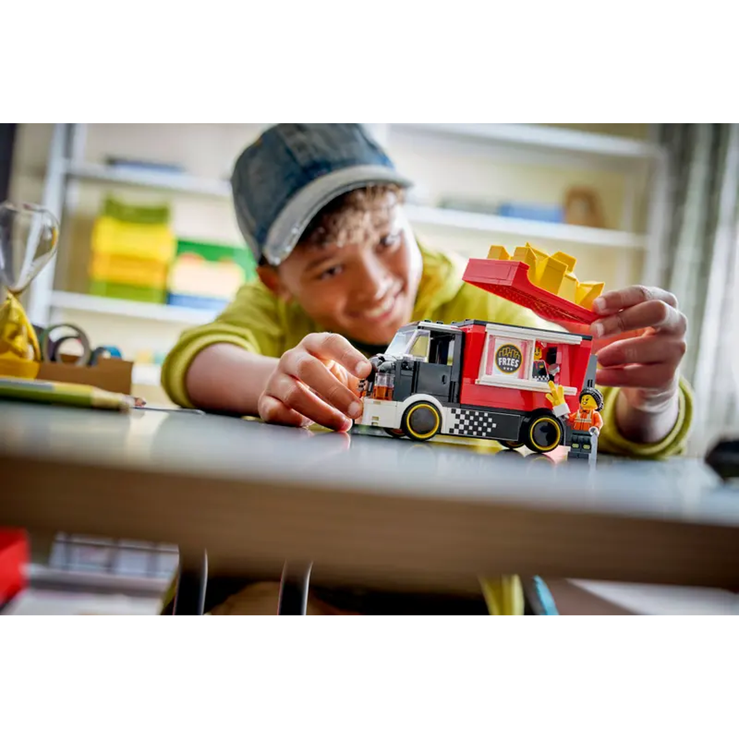 A boy plays with a Lego food truck. The red truck has a fries container. A Lego person stands near the truck.