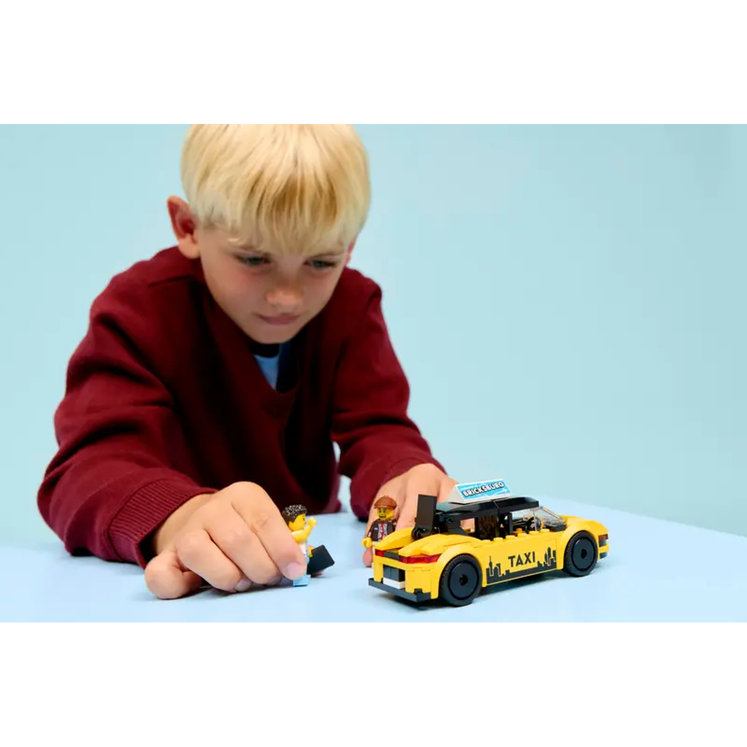 A boy in red sweater plays with a yellow taxi toy and lego figures. The taxi has black wheels and a 'TAXI' sign.