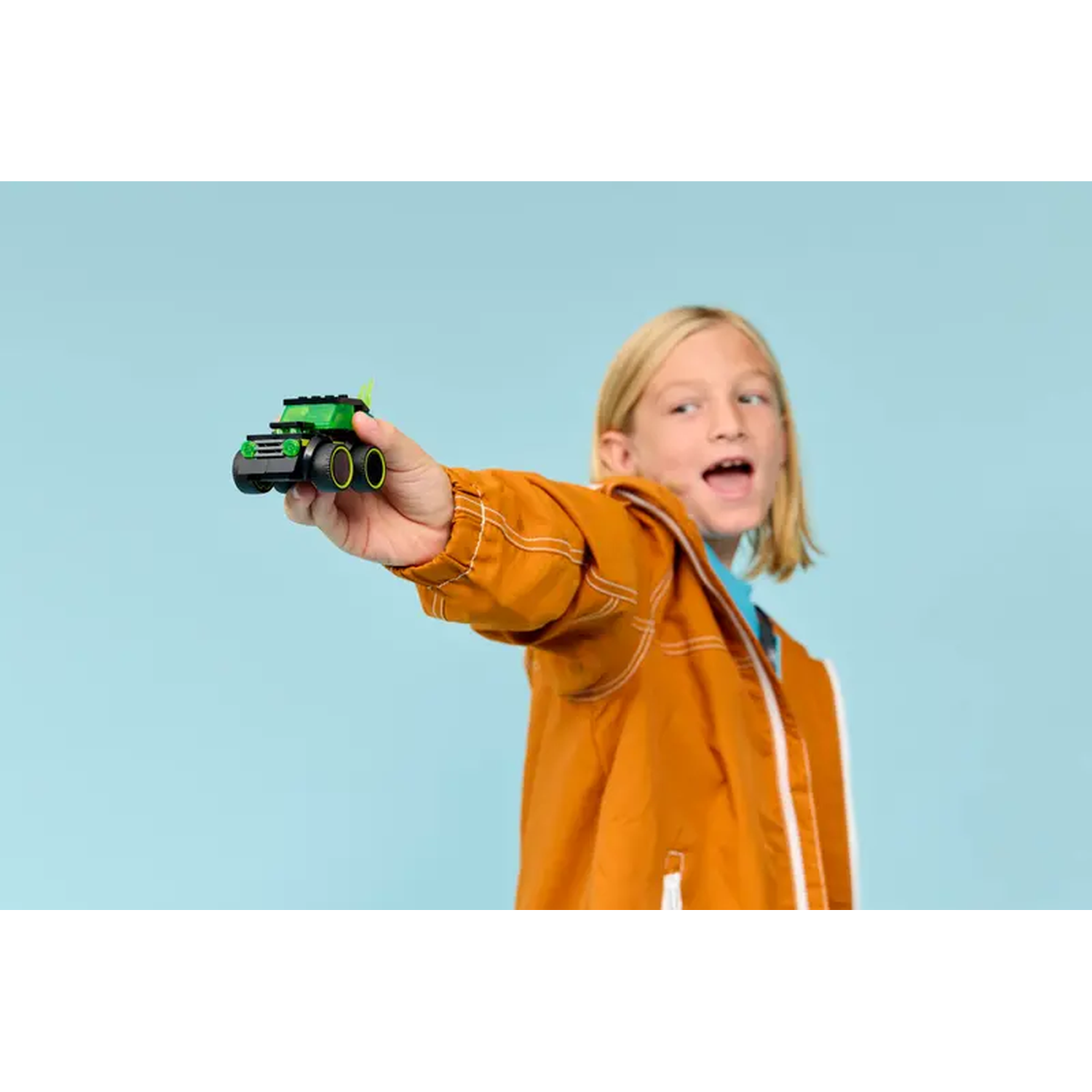 A boy holds a toy car. The car is black and green, and the boy wears an orange jacket against a blue background.