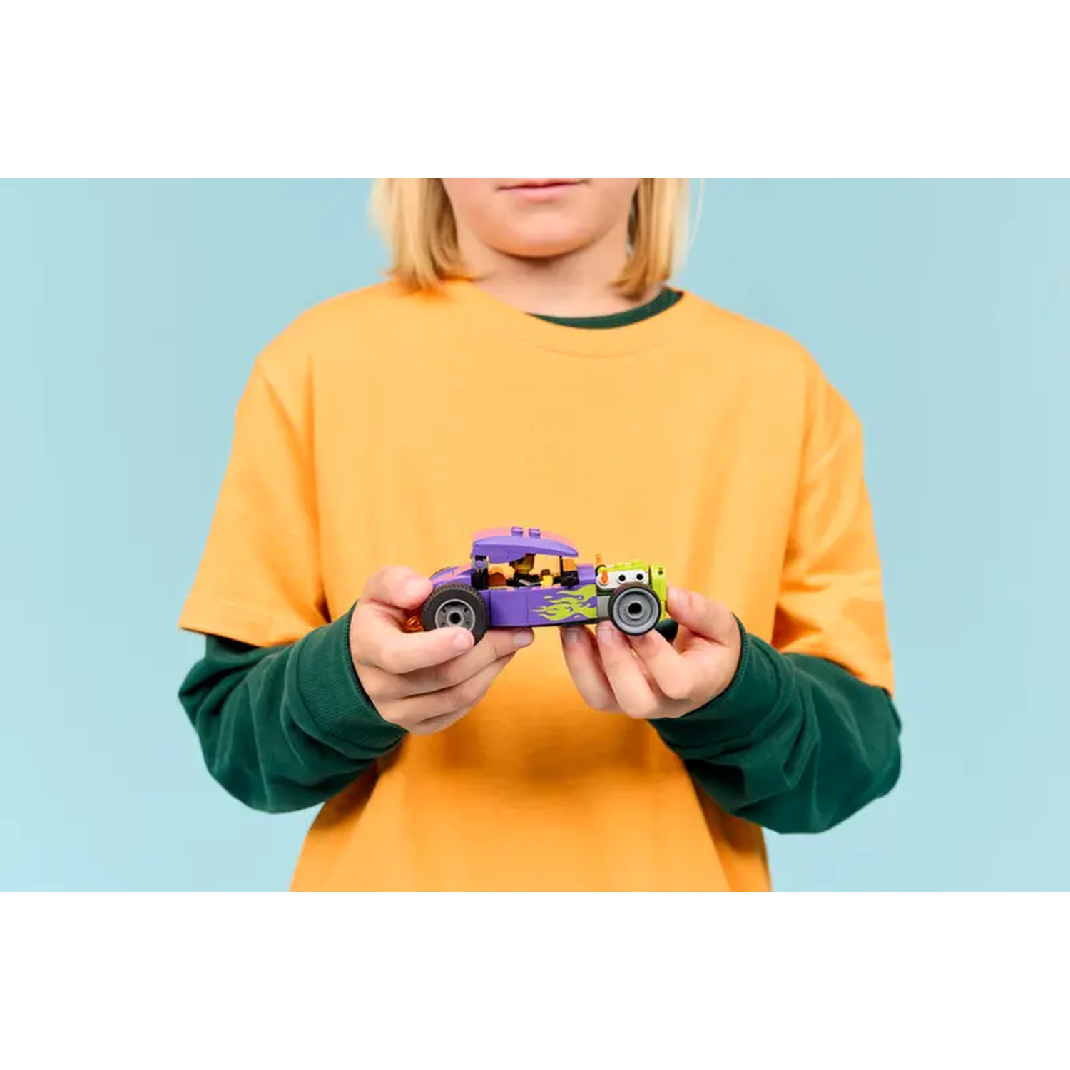 A child holds a purple toy car with green and yellow accents. The child wears an orange shirt with green sleeves, against a blue background.