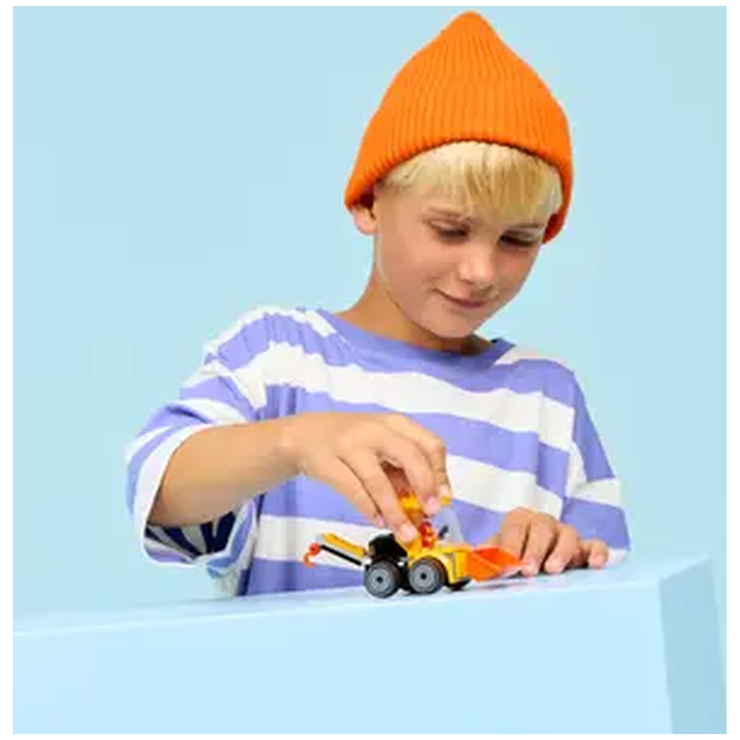 A boy wearing an orange beanie and striped shirt plays with a small toy construction vehicle on a light blue surface.