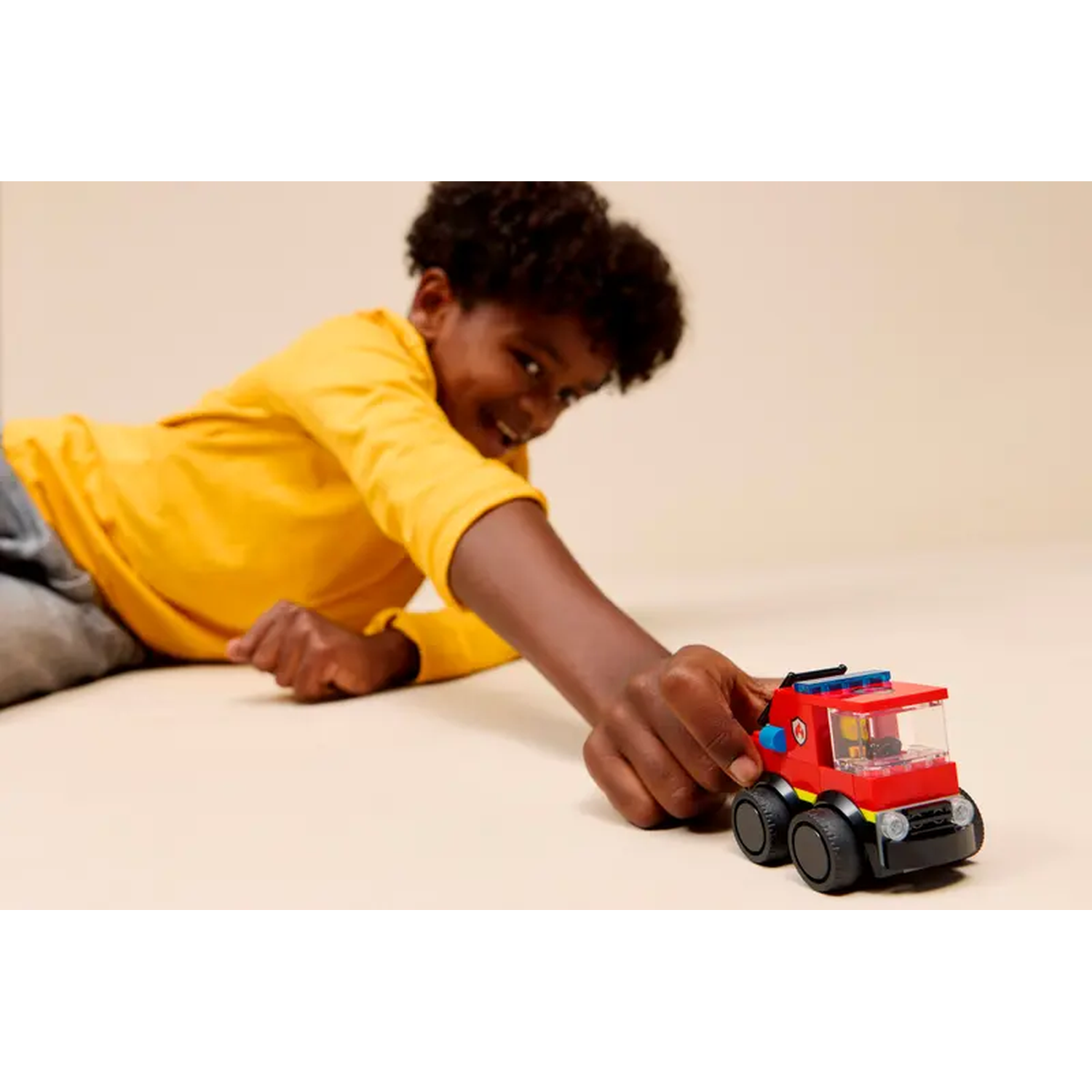 A smiling boy in yellow shirt plays with a red Lego fire truck. The truck has black wheels. Beige background.