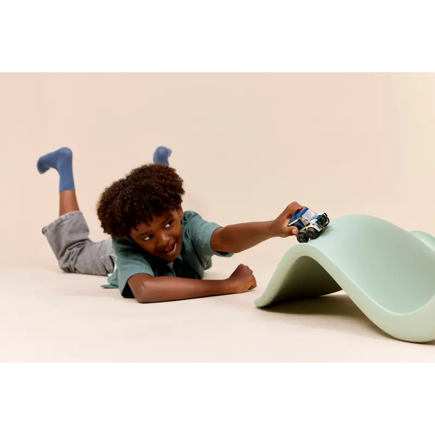 A boy lies on the floor playing with a toy car on a green curved surface. The boy is smiling. Background is light.