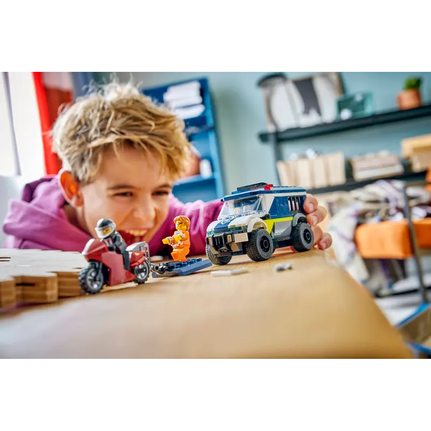 A boy smiles, playing with Lego police car, motorcycle, and figures. The scene is set on a table with a blurred background.