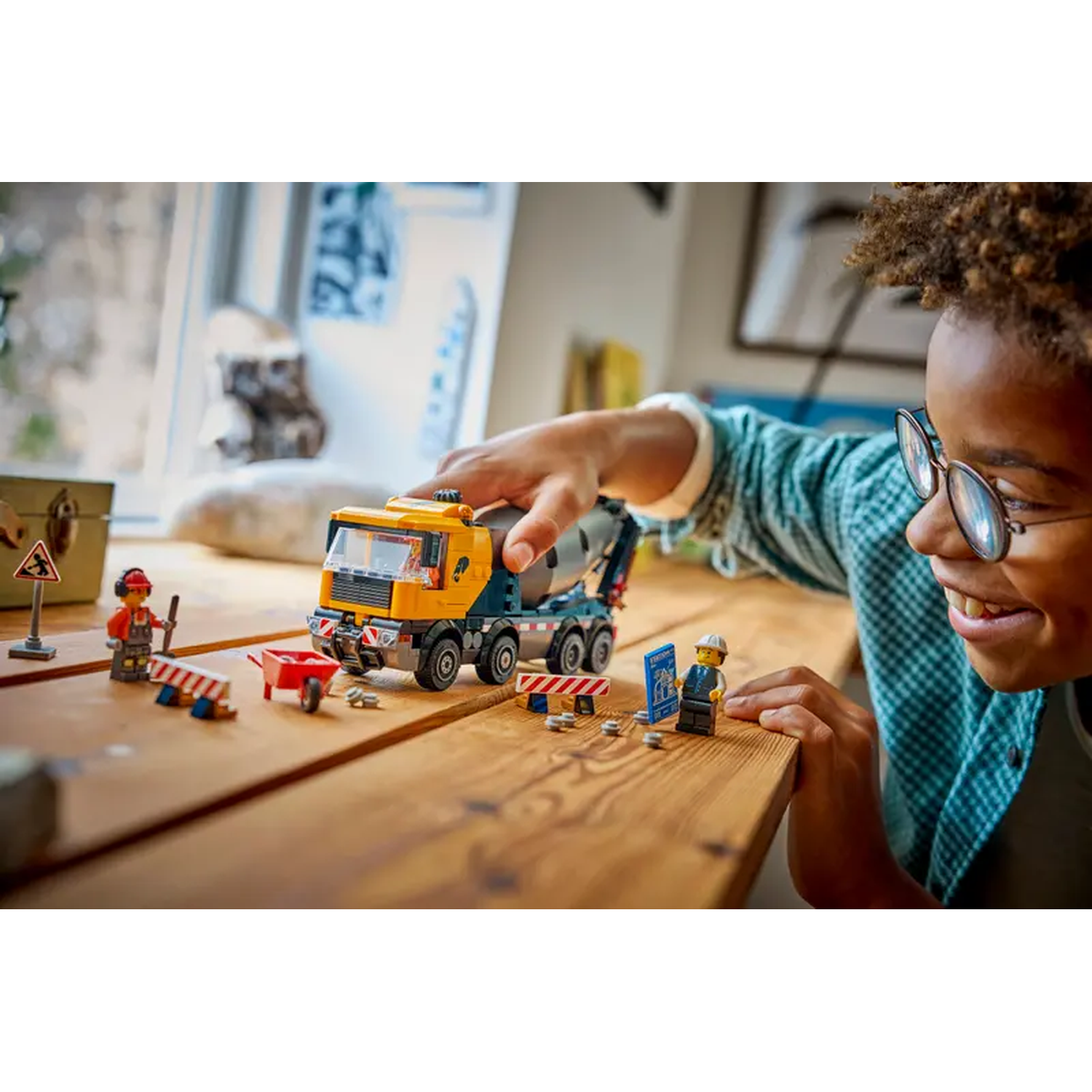 A smiling boy with glasses plays with a Lego construction set on a wooden table. The set includes a cement truck and figures.
