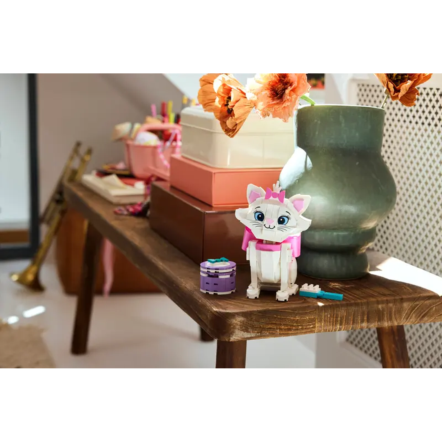 A Lego Marie cat sits on a wooden bench. Behind it are flowers in a vase, and boxes. A trumpet and other items sit nearby.