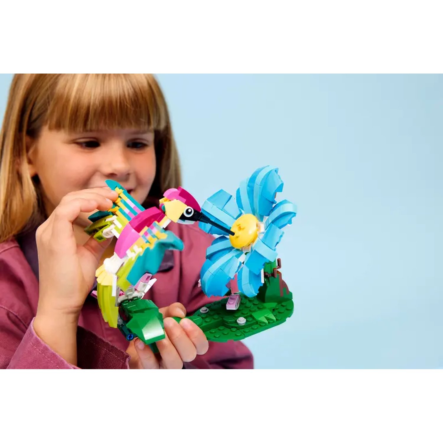 A girl holds a Lego hummingbird and flower. The bird is colorful. The flower is blue with a yellow center. Green base.