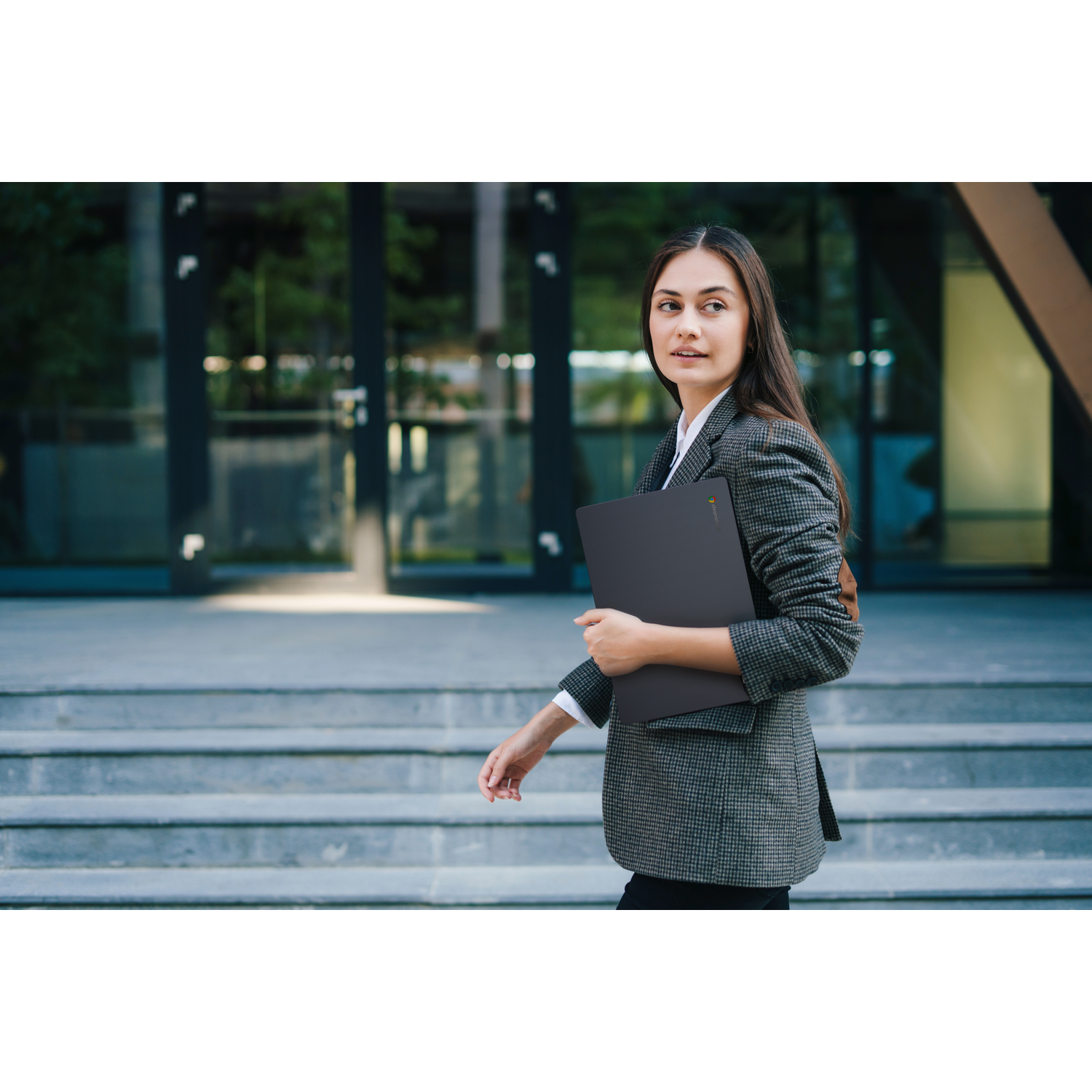 A woman in a blazer walks, holding a laptop. She is outside, with a modern building and steps in the background.