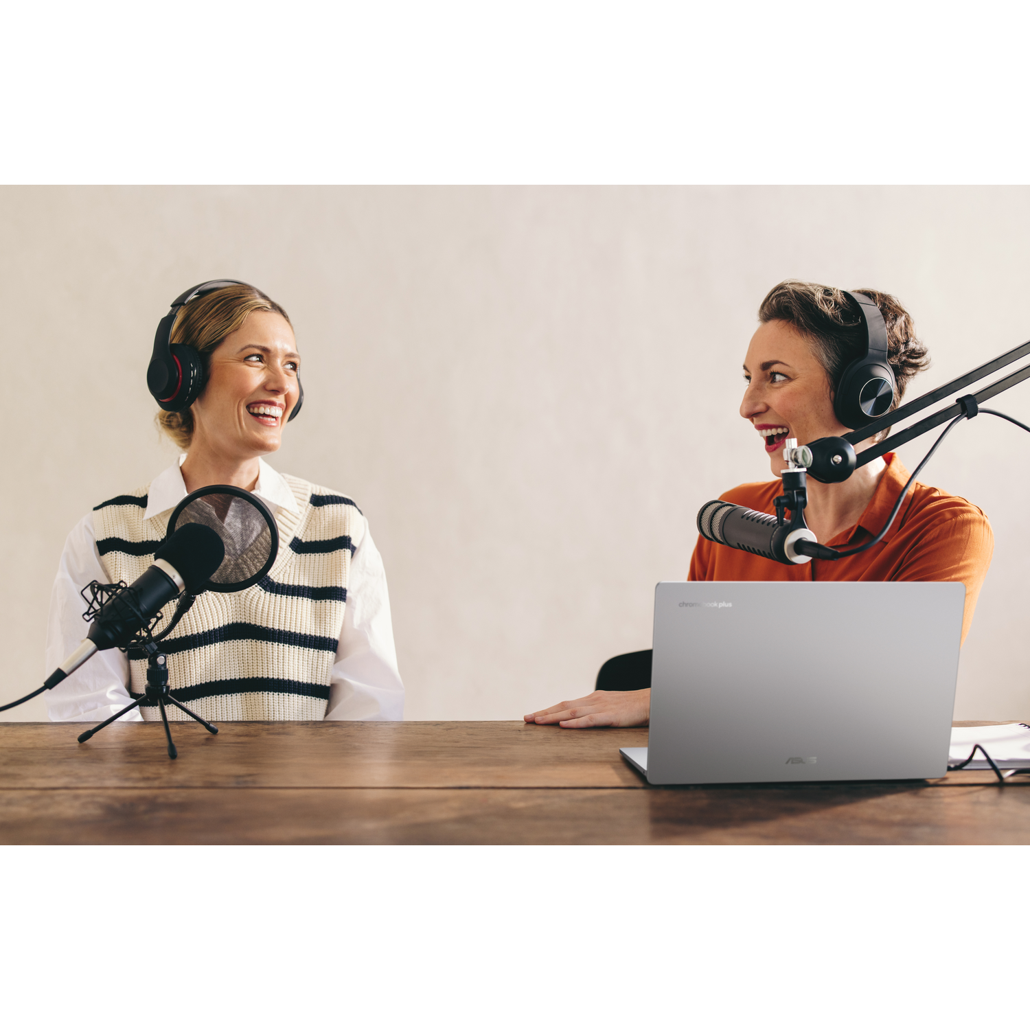 Two women with headphones and microphones at a table, smiling. A laptop is in the foreground. Background is a neutral wall.