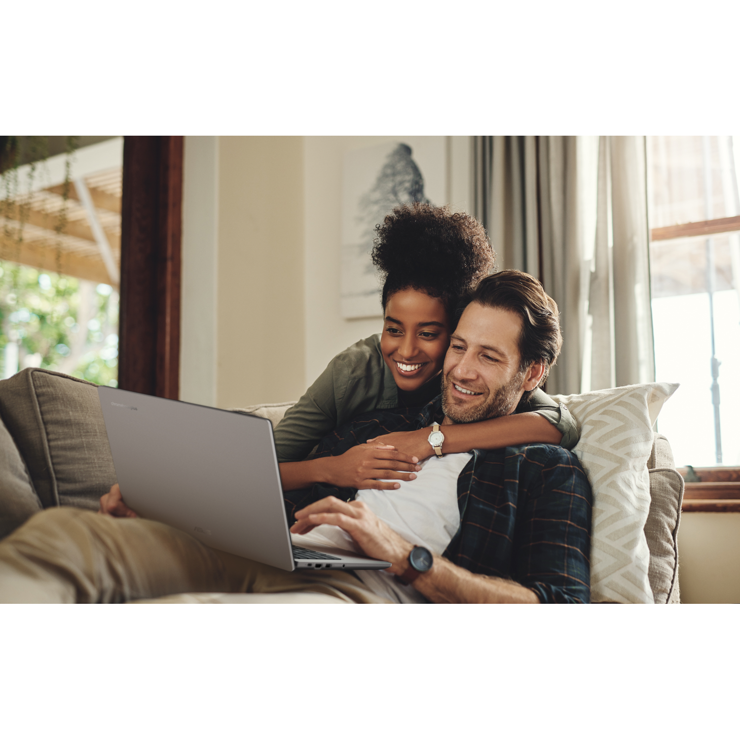 A smiling couple on a couch, looking at a laptop. Woman hugs man from behind. Bright room with window.