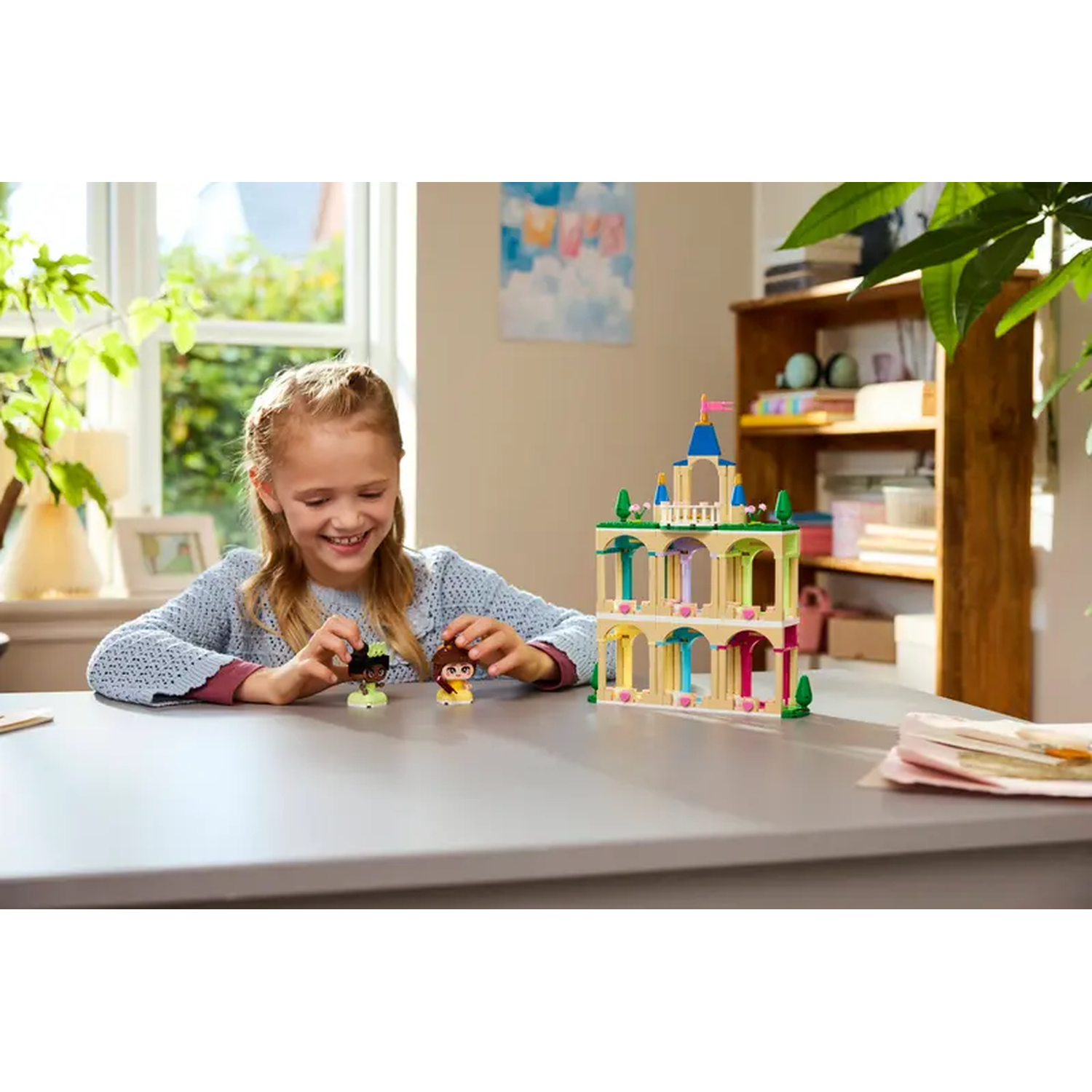 A girl smiles, playing with Lego figures and a castle. A wooden bookshelf and window are in the background.