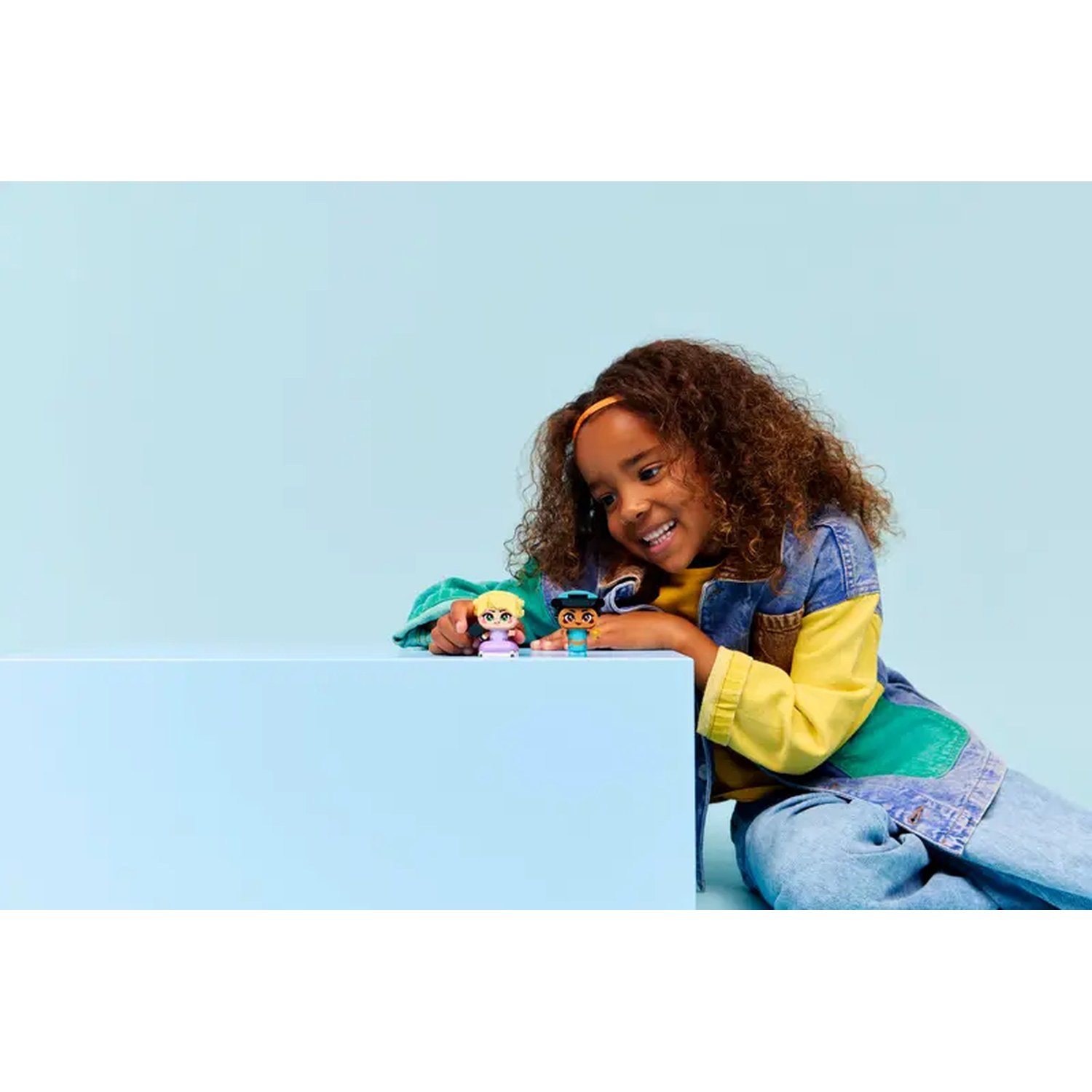 A young girl smiles, leaning on a light blue block. She has two small toy figures. The background is also light blue.