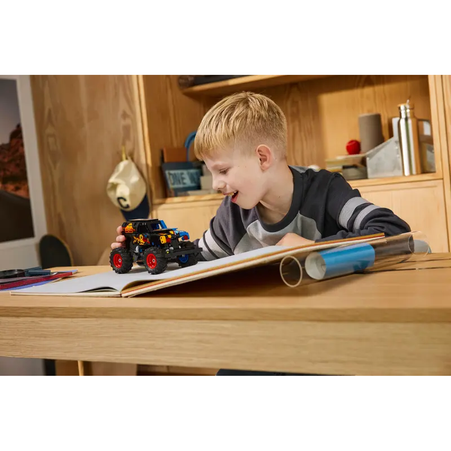 A boy smiles, looking at a toy monster truck on a book. Wooden table, background shelves with items and a hat.