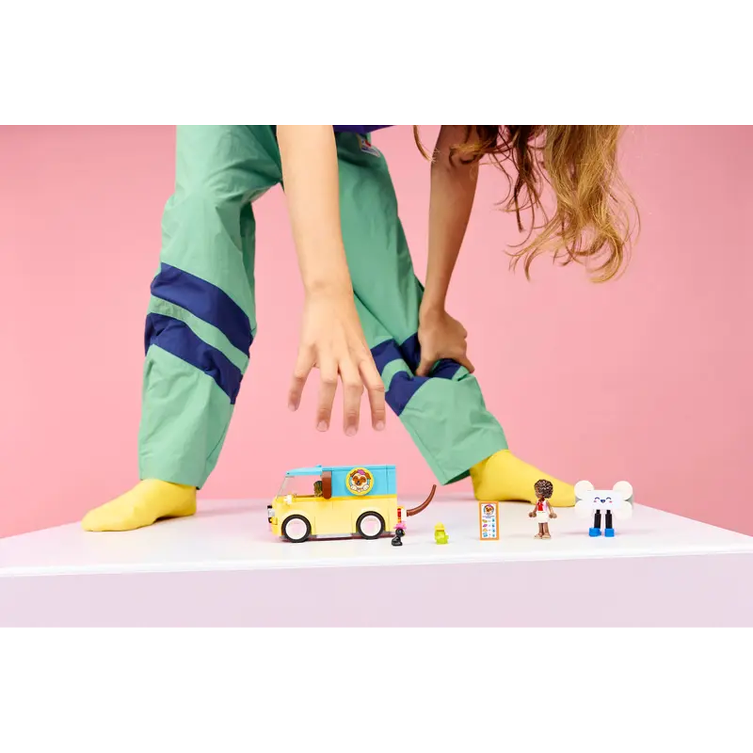 A person in green pants with blue stripes and yellow socks. A yellow van toy is on a white table with other toys. Pink background.