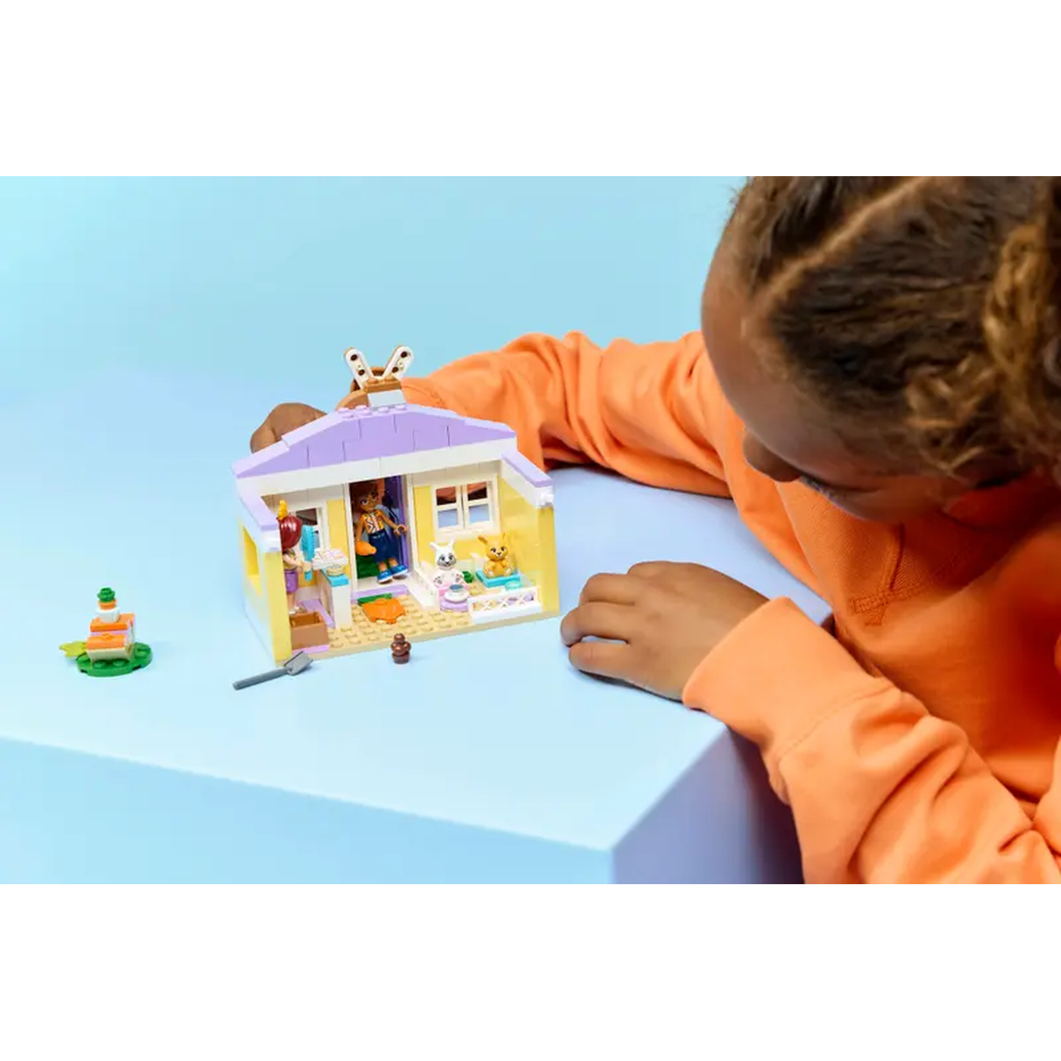A child playing with a Lego house. The house is yellow and purple, with small figures inside. A small cake is on the side.