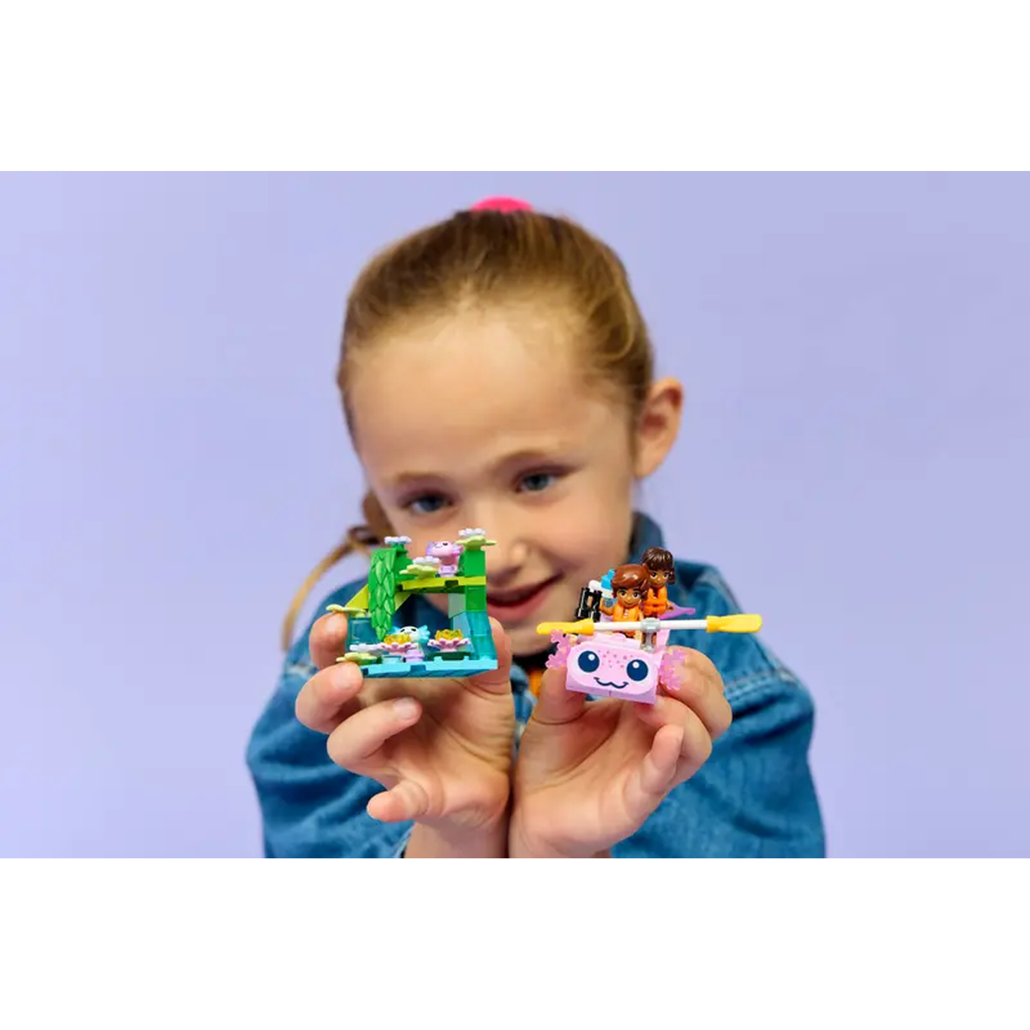 Een meisje houdt twee Lego sets vast: een groene structuur en een roze, axolotl-vormige boot met Lego figuren, tegen een paarse achtergrond.