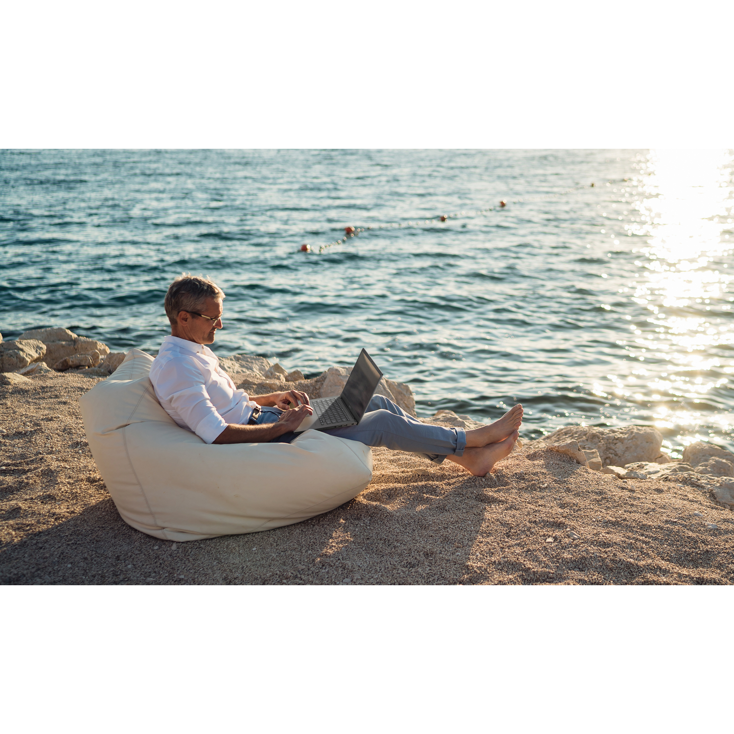 Man zit op een zitzak op het strand, werkend op een laptop. De zee en rotsen zijn op de achtergrond. Het is een zonnige dag.