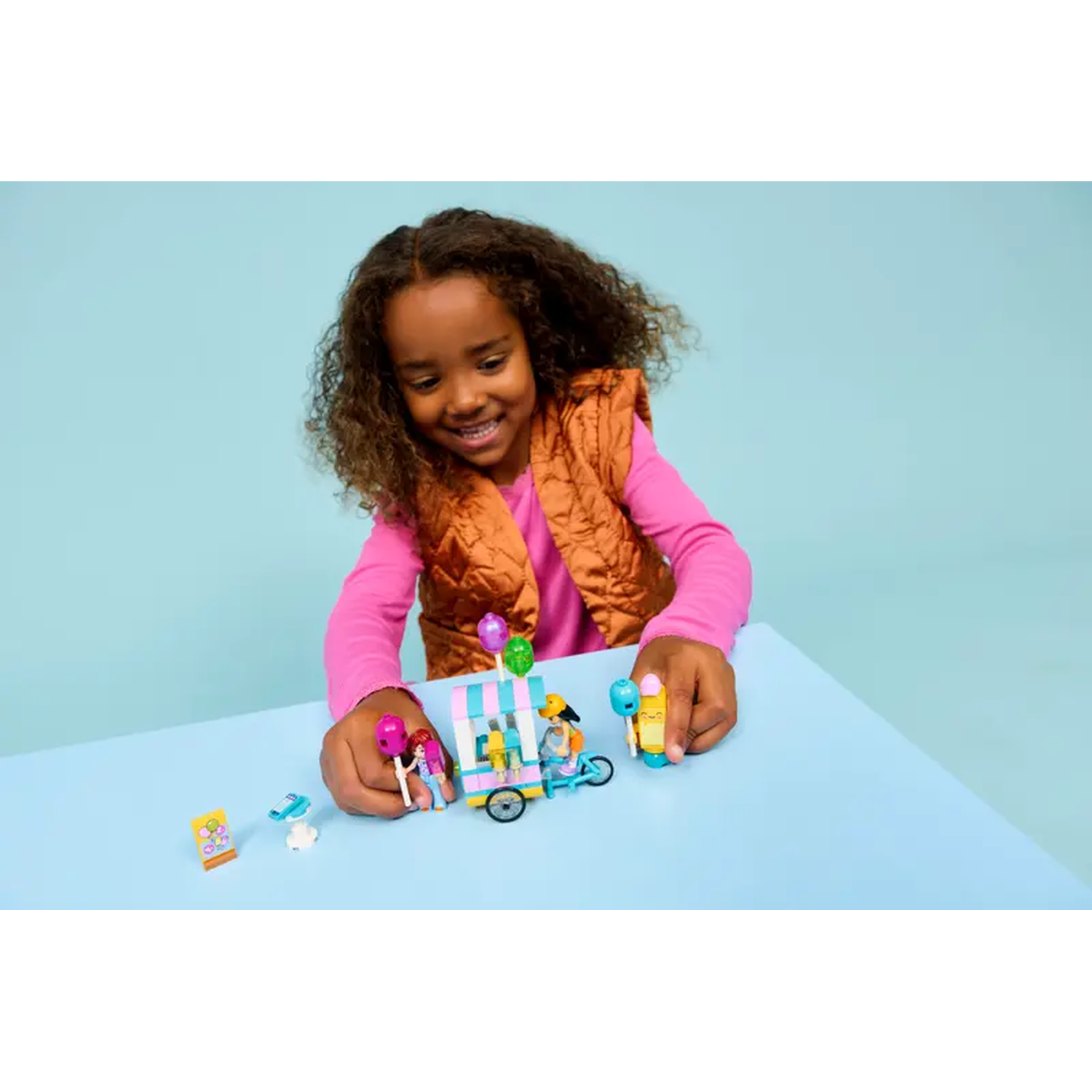 Een jong meisje lacht en speelt met Lego-speelgoed, een klein karretje met ballonnen en andere kleurrijke Lego-stukken op een blauwe tafel.