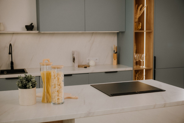 Nowoczesna kuchnia z blatem, makaronem w słoikach, rośliną i płytą grzewczą. Szafki są niebiesko-szare.