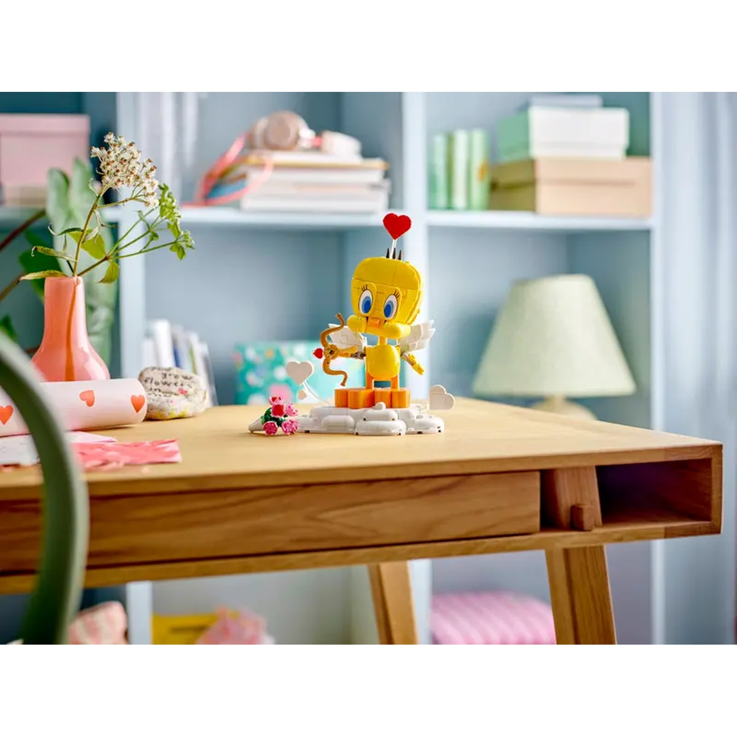 Un Lego de Titi avec un cœur sur la tête, tenant un arc et une flèche, est sur un bureau en bois. Des fleurs et une lampe sont en arrière-plan.