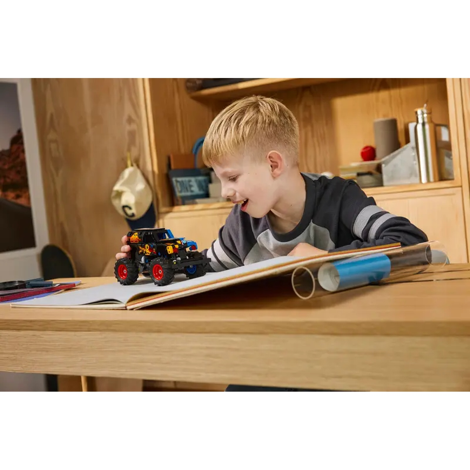 A boy smiles at a toy truck on a book. The scene includes a wooden table, and objects in the background.