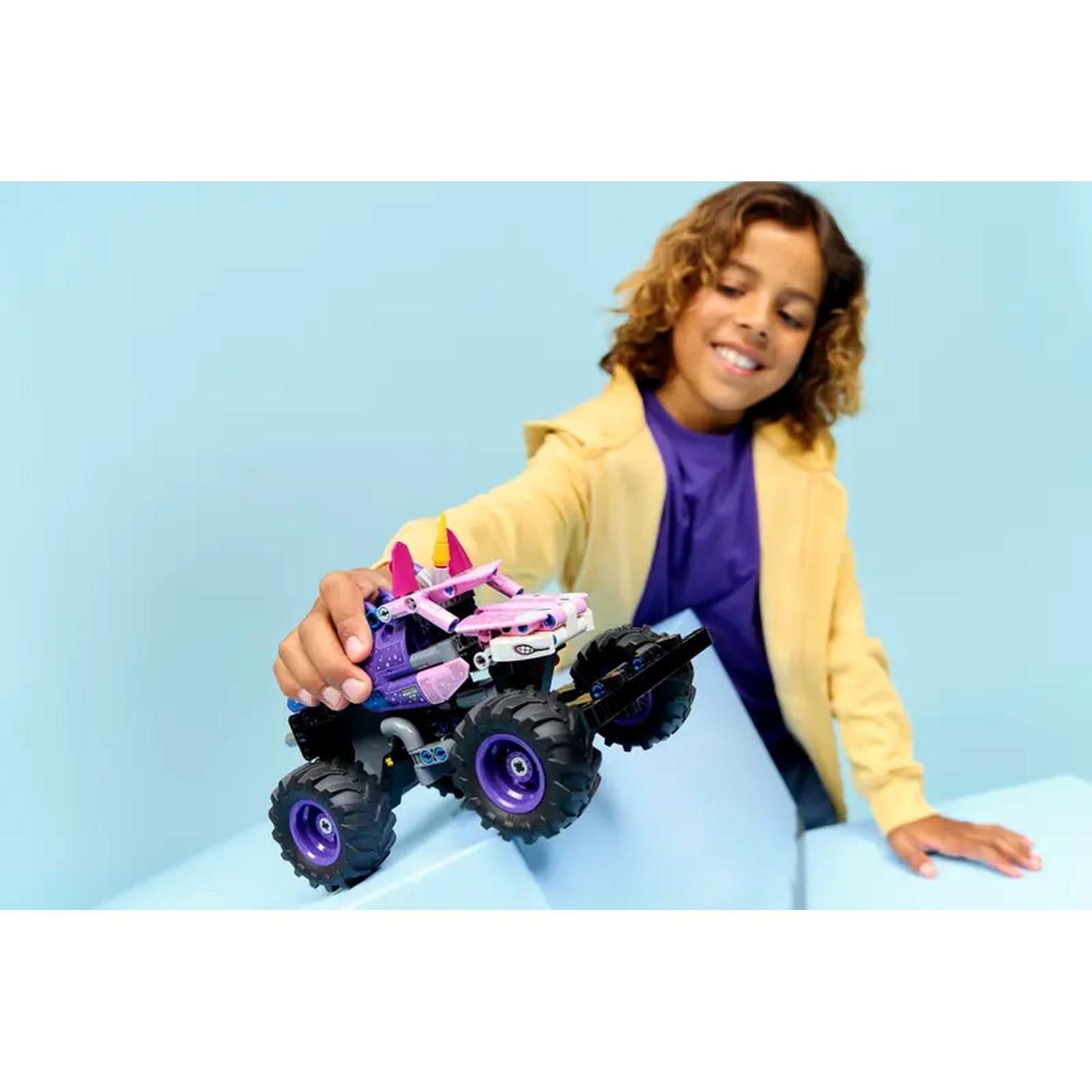 A smiling boy in a yellow jacket holds a toy monster truck. The truck is purple, pink, and black, with large wheels.