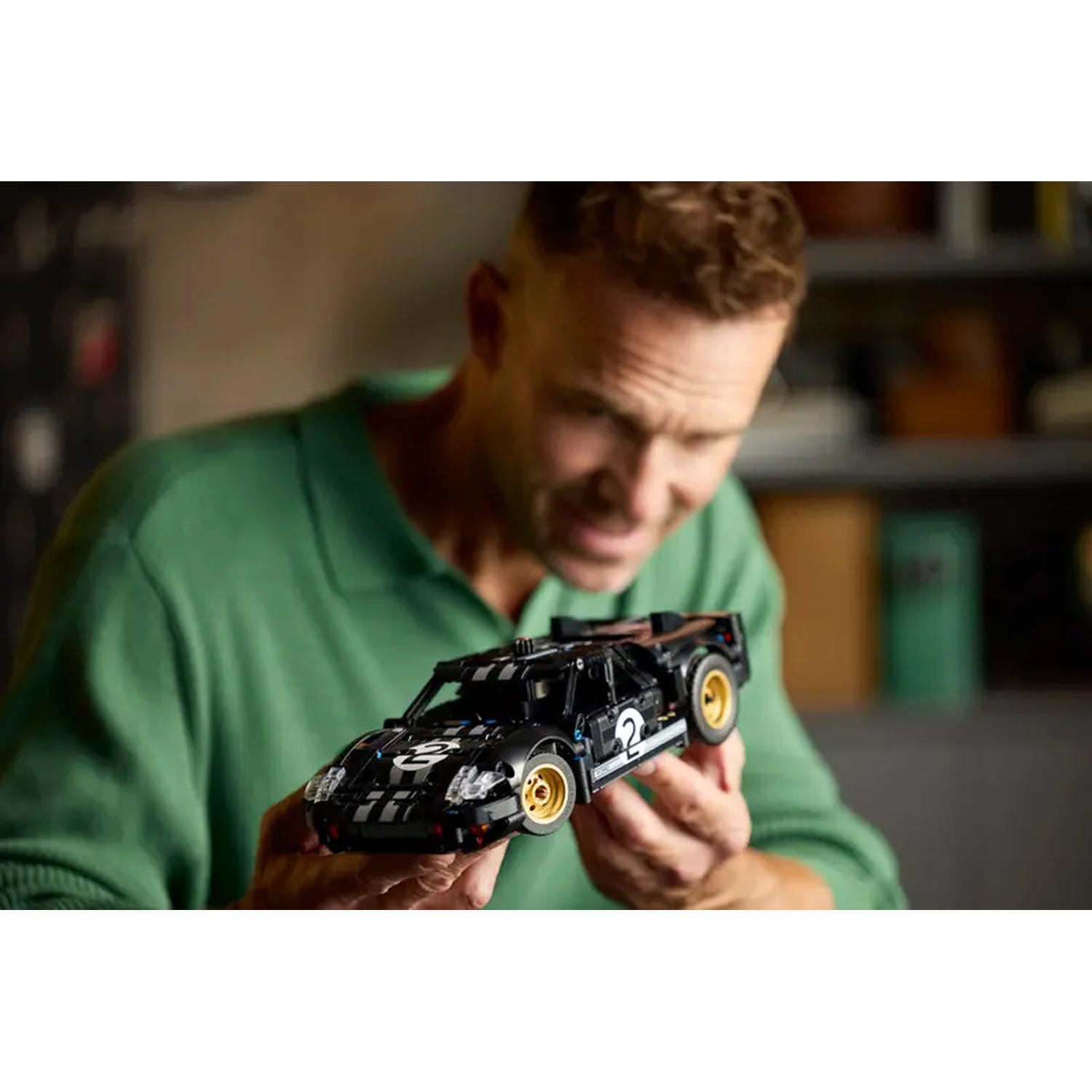 A man holds a black toy race car with gold wheels. The car has the number 2 on it. The man wears a green shirt.