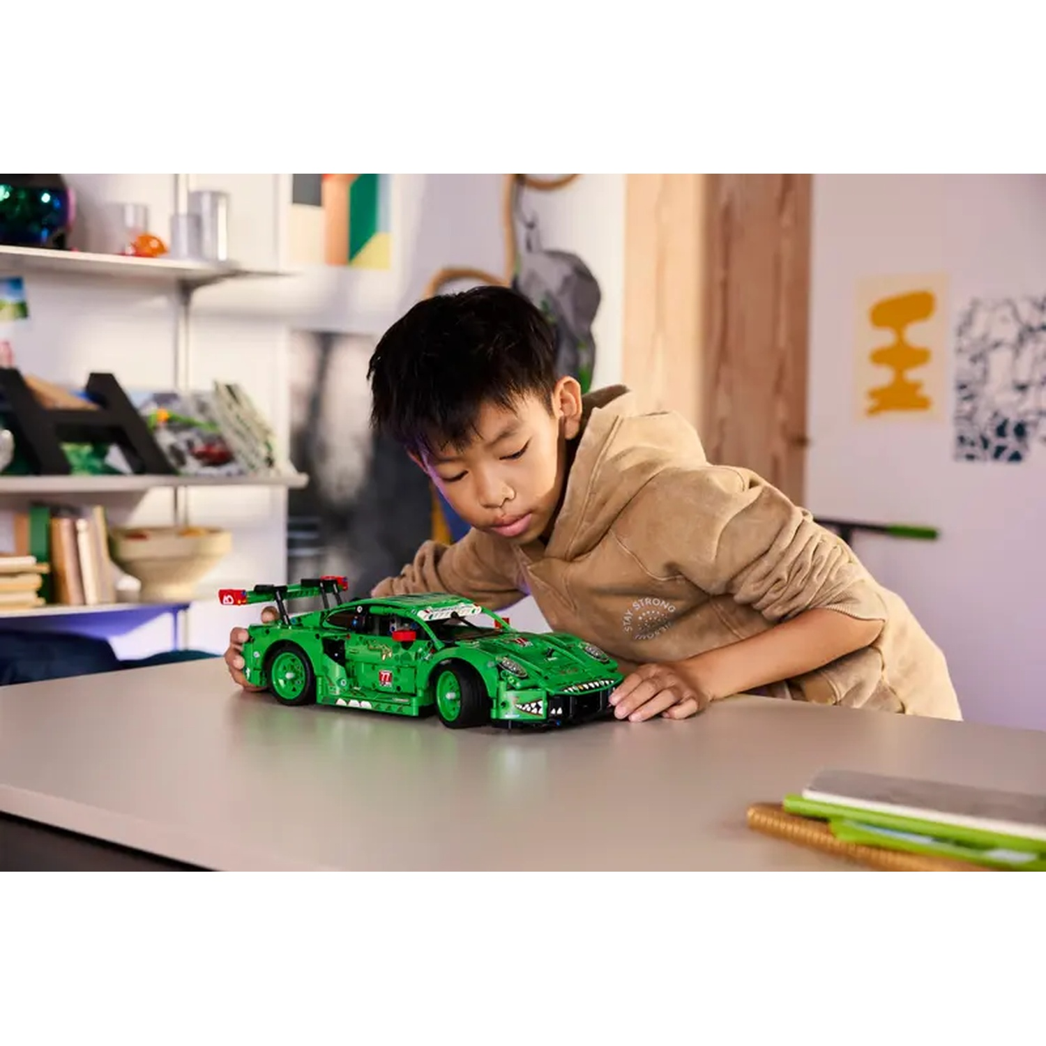 A young boy examines a green Lego race car on a table. The car sits on a gray surface. Background includes shelves and art.