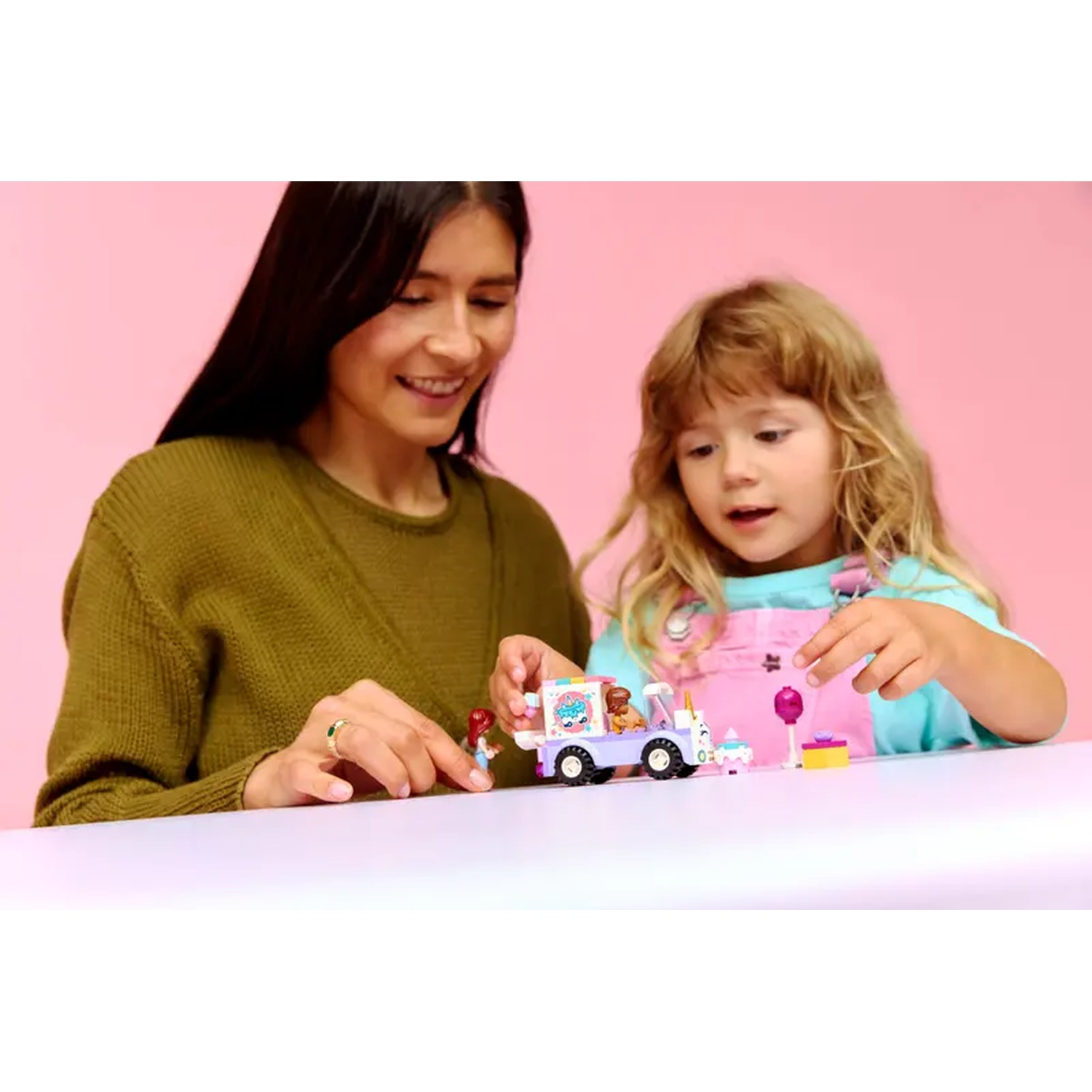 A woman and a girl play with a toy ice cream truck. The truck is white and purple. The background is pink.