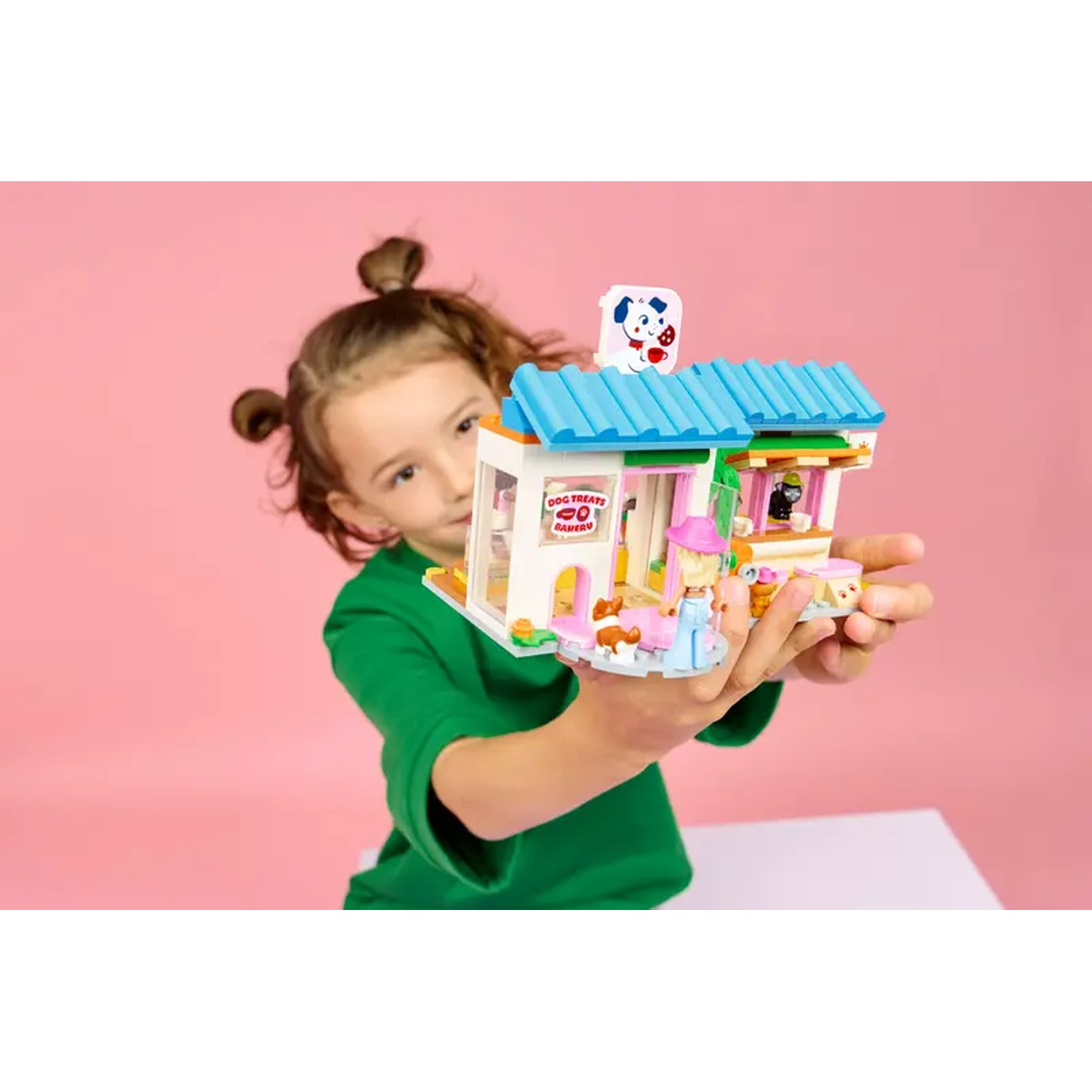 A girl holds a toy dog bakery. The bakery has a blue roof, white walls, and a pink background. A dog figure is on top.