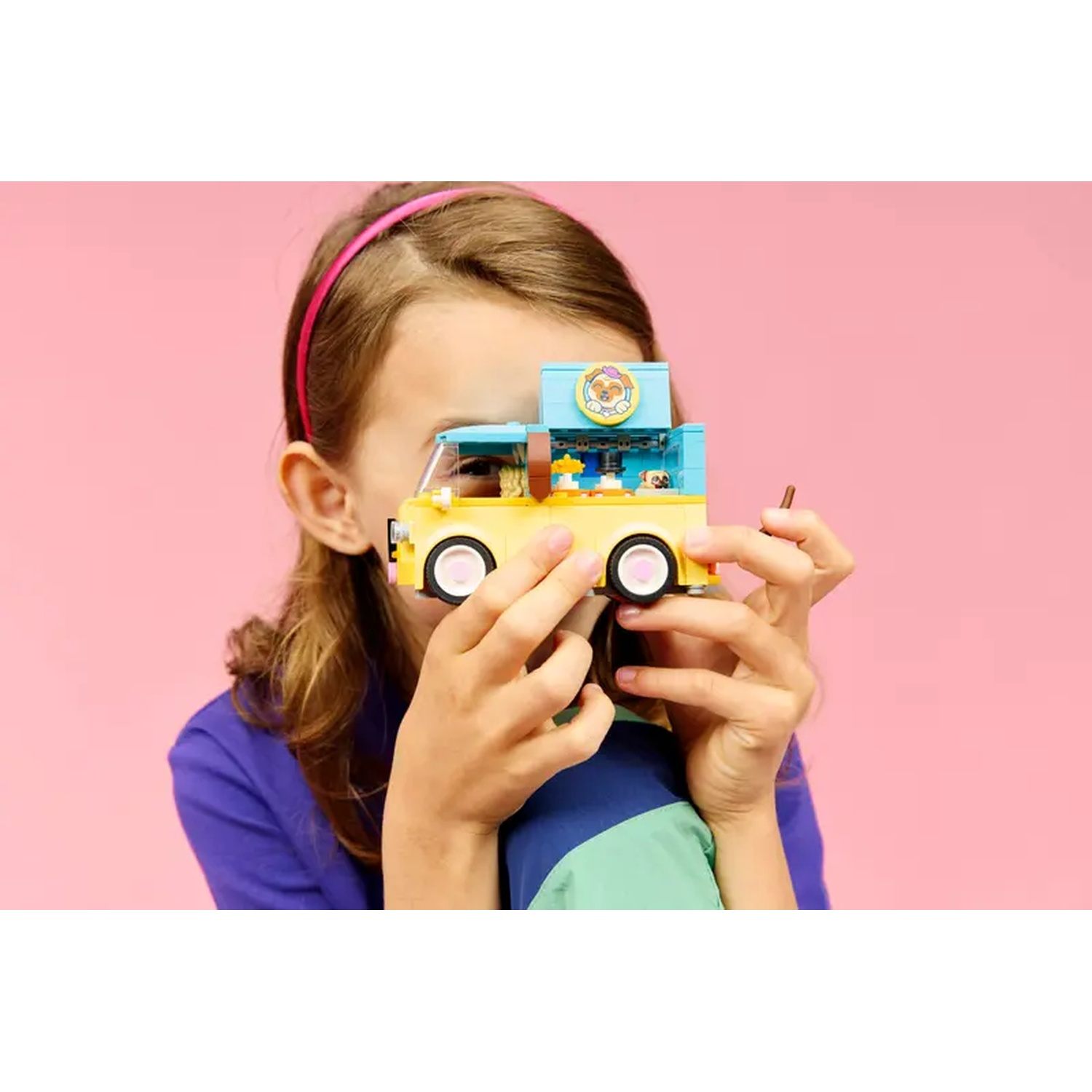A girl holds a yellow toy food truck in front of her face. Pink background. The truck has a blue top and white wheels.