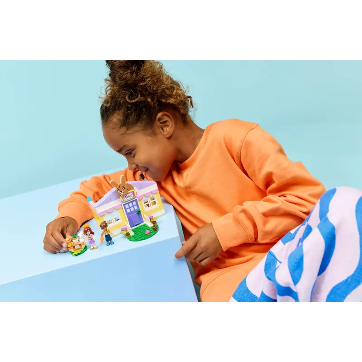 A young girl examines a Lego set of a building with figures and animals. She wears orange and blue striped pants.