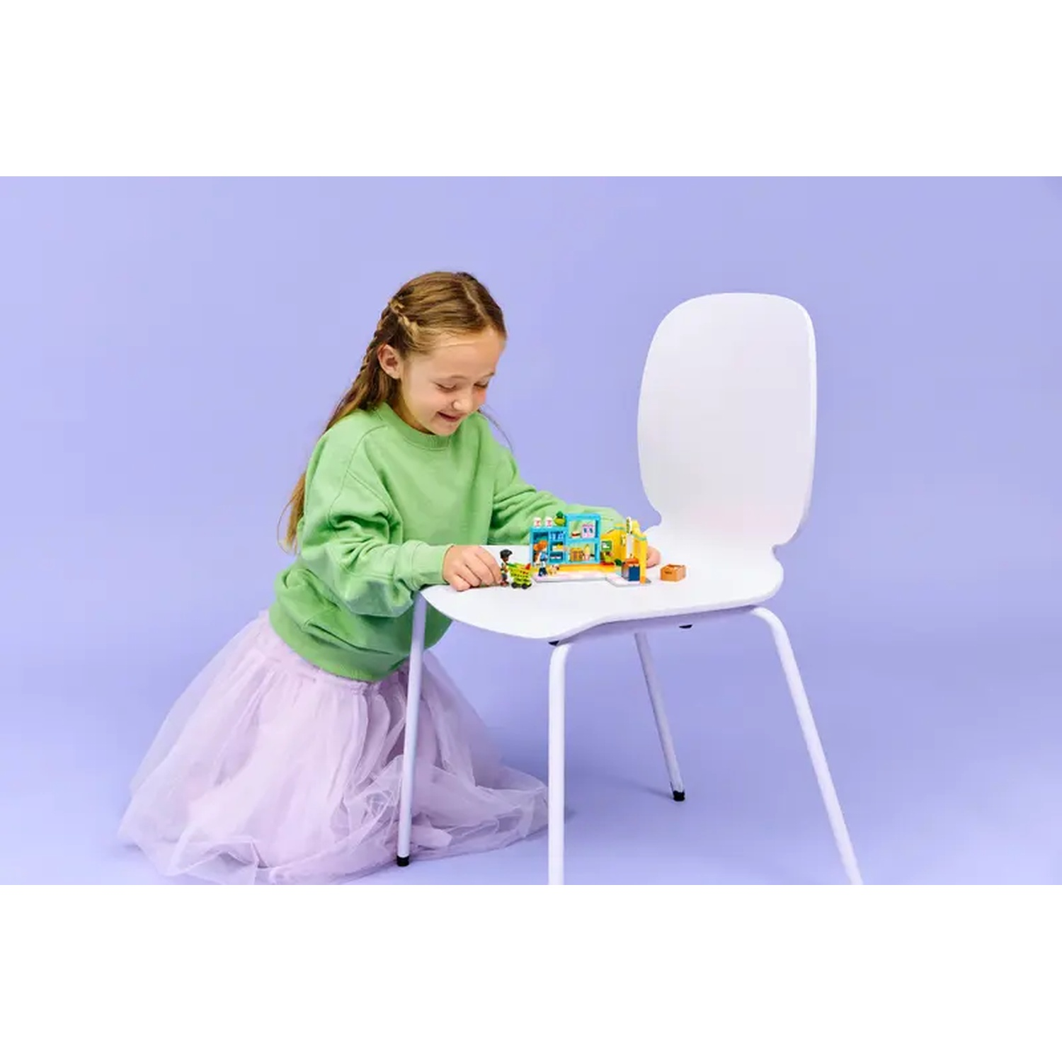 A girl in green sweater and pink skirt plays with building blocks on a white chair. Purple background.