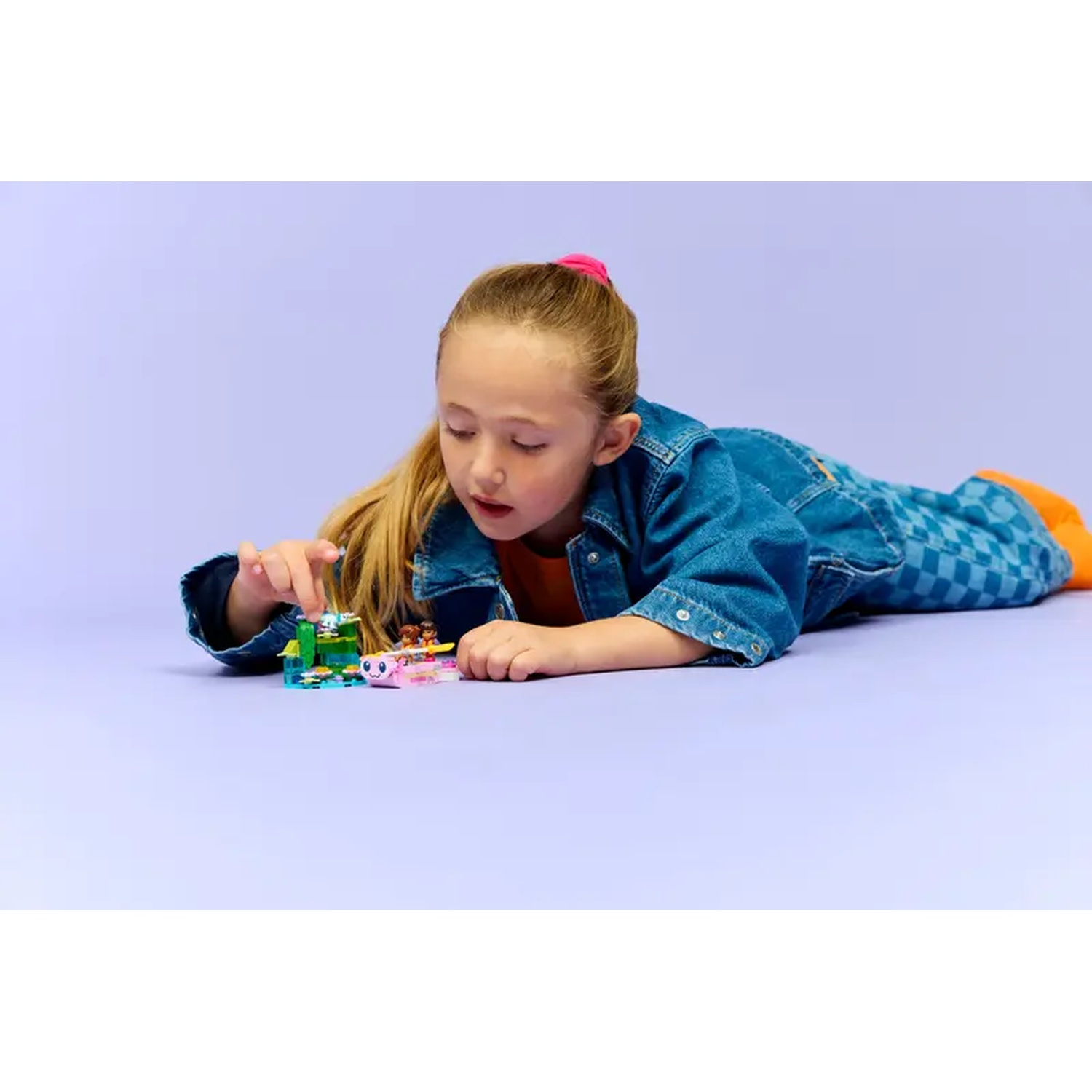 A young girl is lying on a purple surface, playing with colorful toy blocks. She's wearing a denim jacket and jeans.