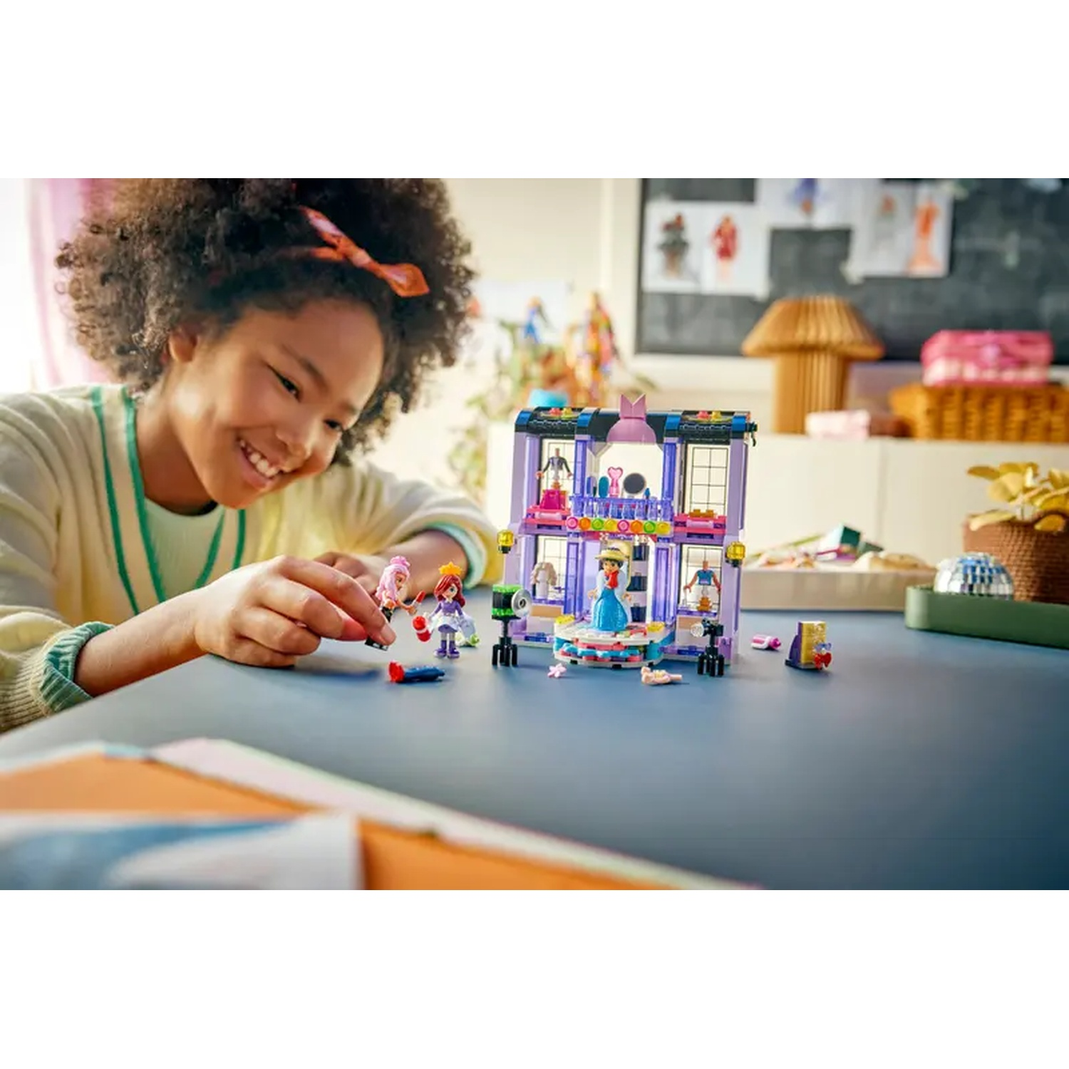 A girl smiles at a LEGO set. The set is a shop with miniature figures. The table is blue. The background is a room.