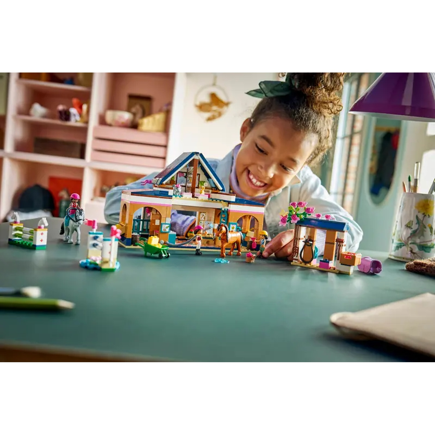 A girl smiles at a Lego horse stable set. The set has horses, figures, and buildings. A horse and rider are on the left.