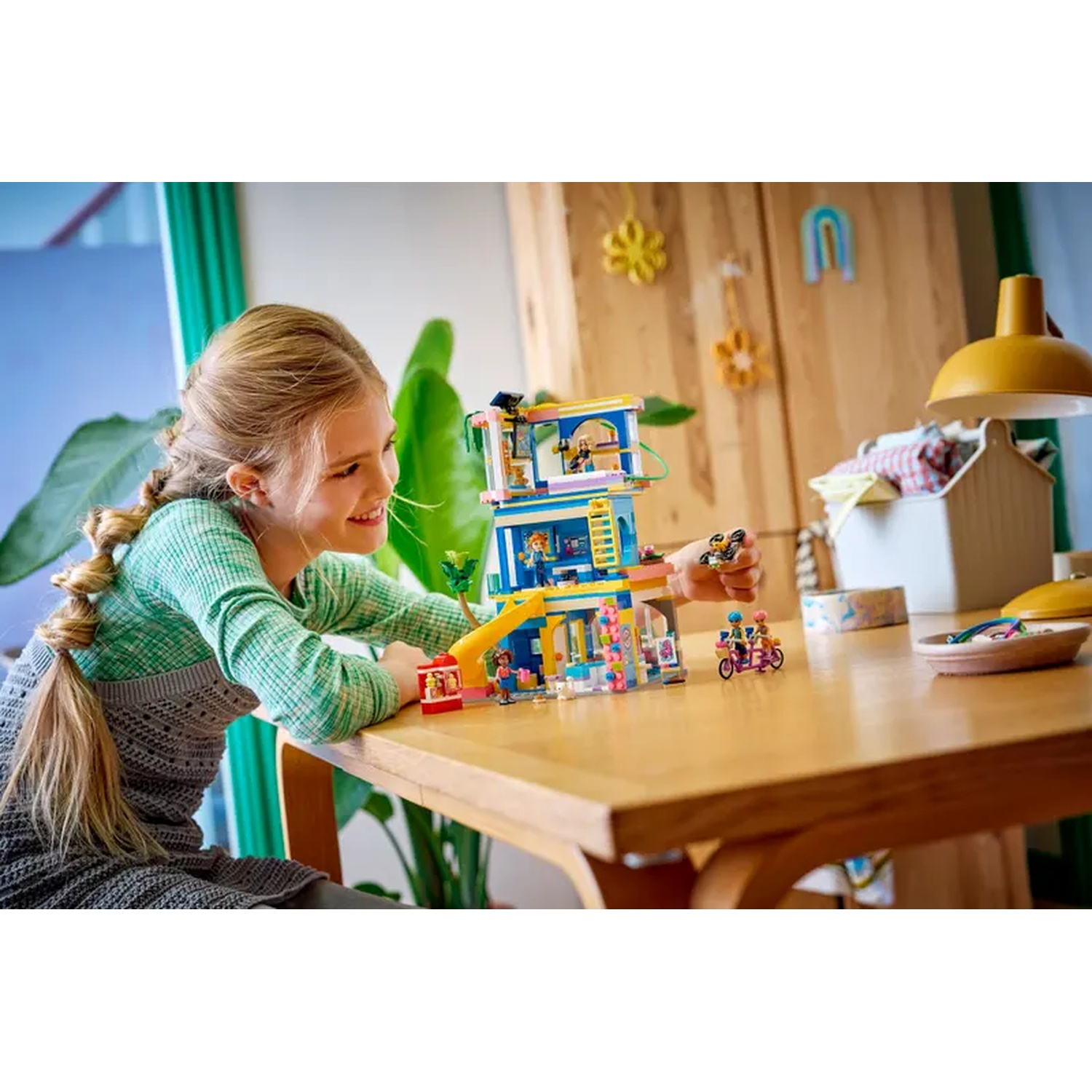 A girl plays with Lego. There is a colorful Lego house, and two Lego figures on a bike. The background has a wooden cabinet.