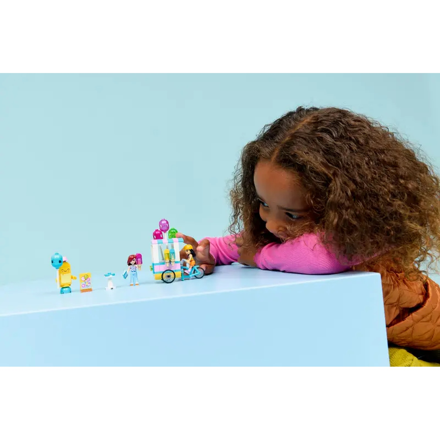 A girl looks at Lego toys. There is an ice cream cart, figures, and other small toy figures on a blue table. Background is blue.