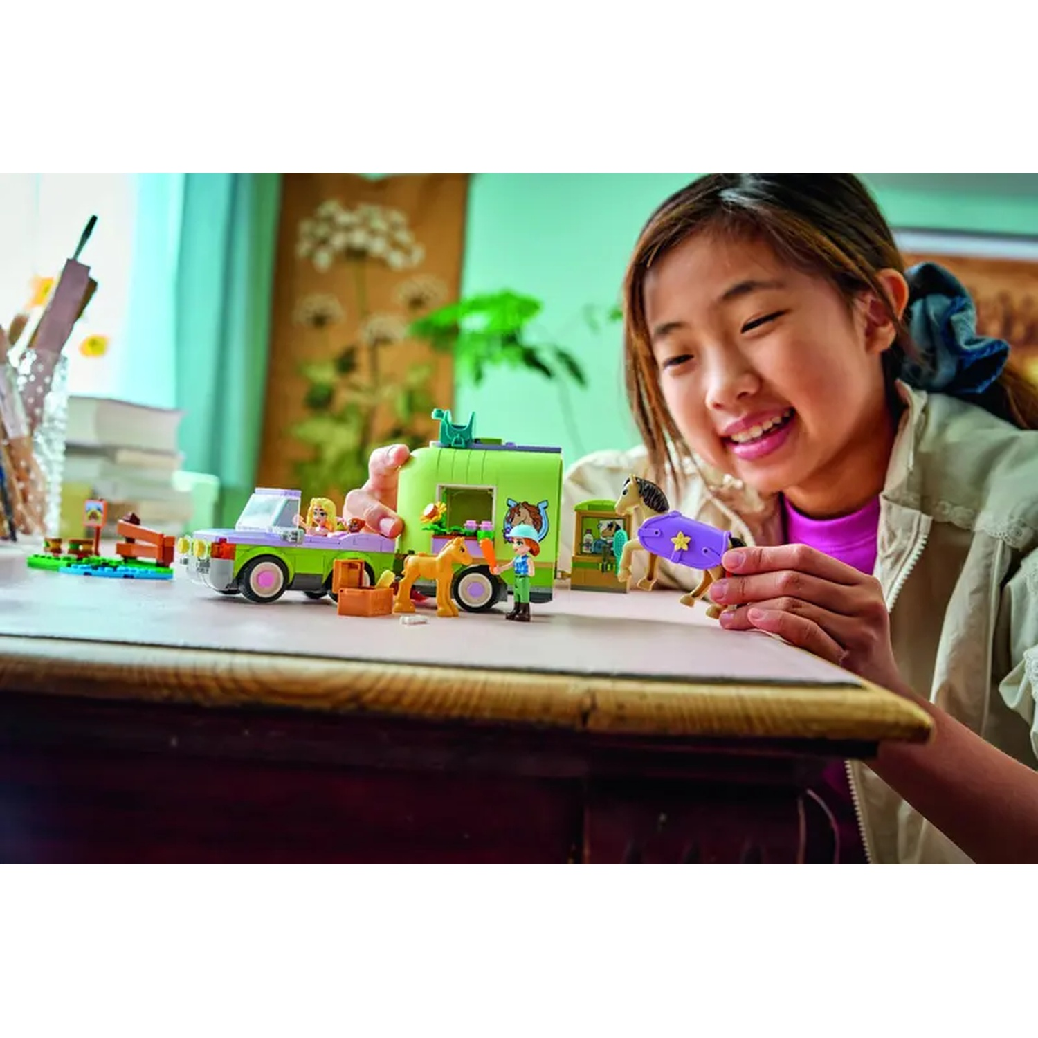 A girl smiles, playing with a toy horse and a LEGO set. The set includes a green truck, a brown horse, and minifigures.