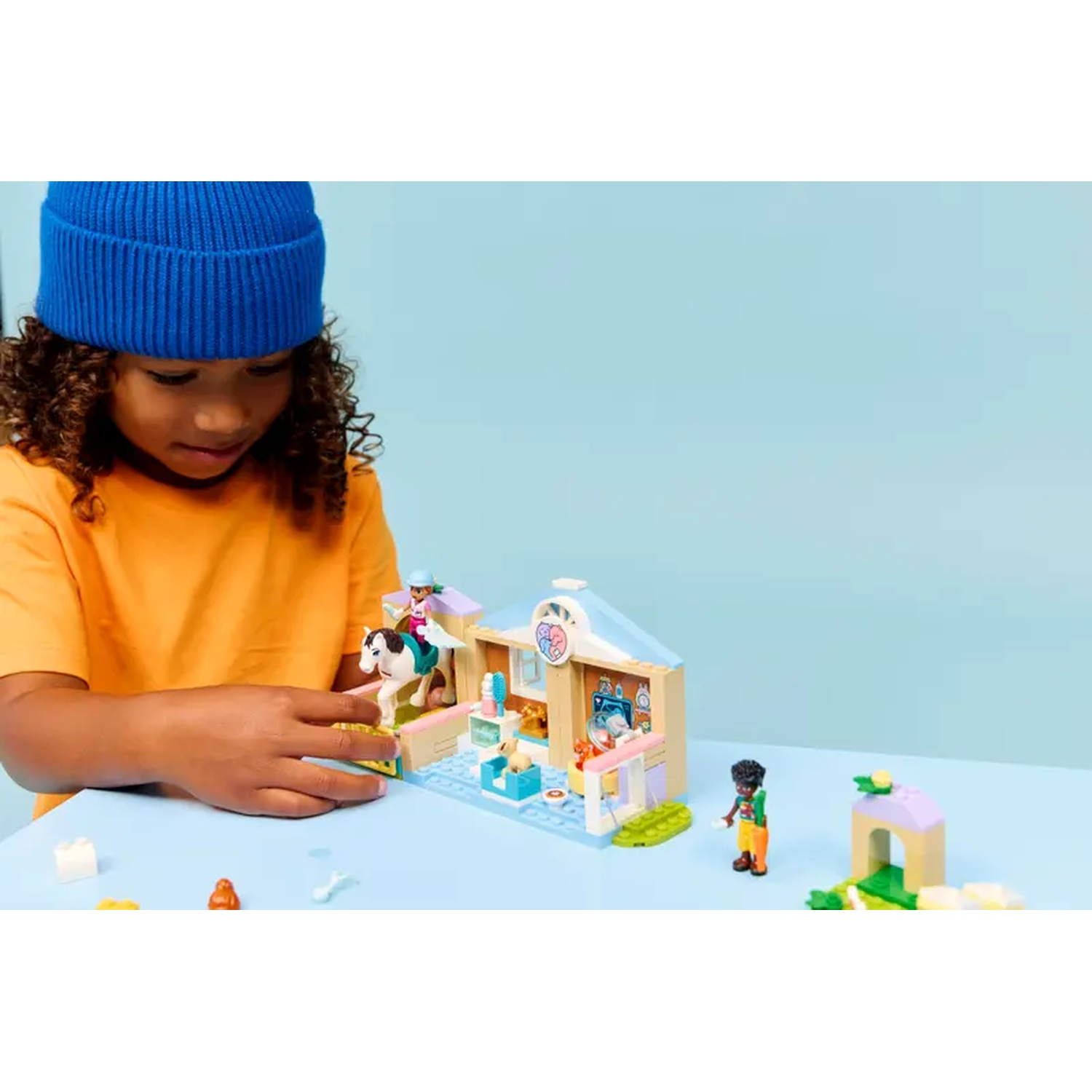 A child in an orange shirt and blue hat plays with a Lego set depicting a vet clinic, with figures and a horse.