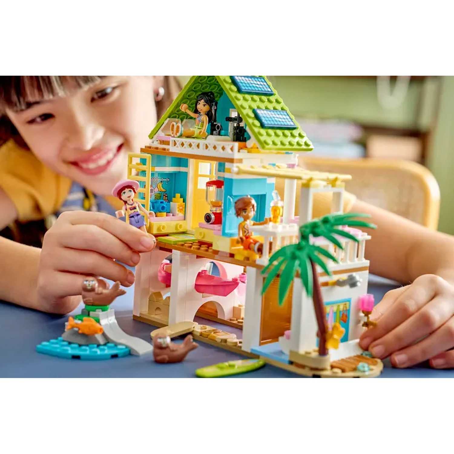 A girl plays with a Lego beach house. It has figures, a palm tree, and solar panels. The girl is smiling.