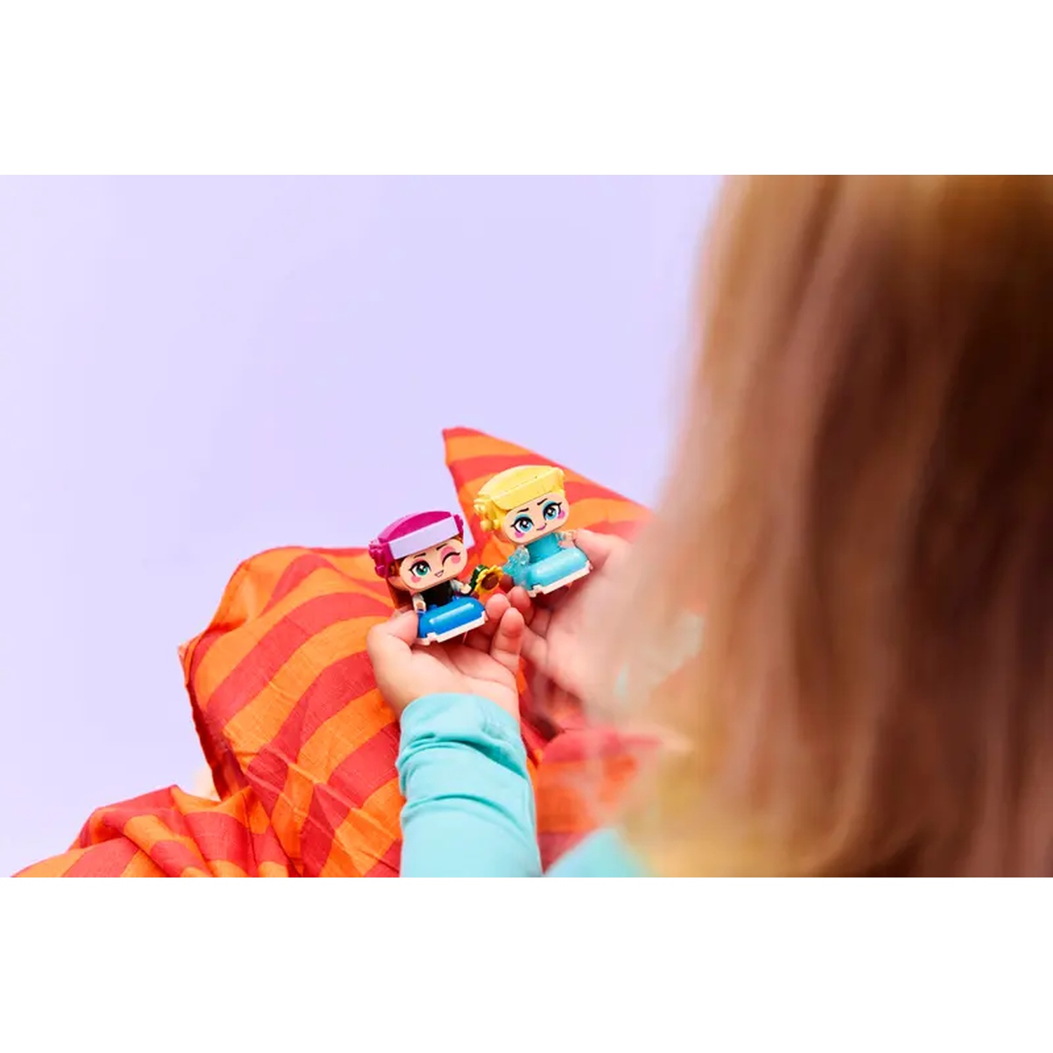 Handen van een kind die twee kleine speelfiguren vasthouden. De figuren zijn stripachtige karakters met kleurrijke outfits. Achtergrond is een rode en oranje sjaal.