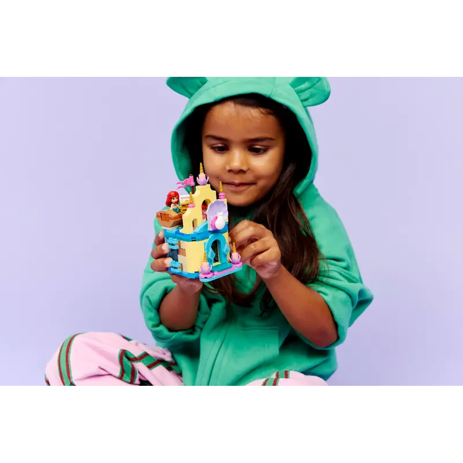 Een meisje in een groene hoodie houdt een Lego kasteel vast. Ze zit op de grond met een roze broek en een paarse achtergrond.