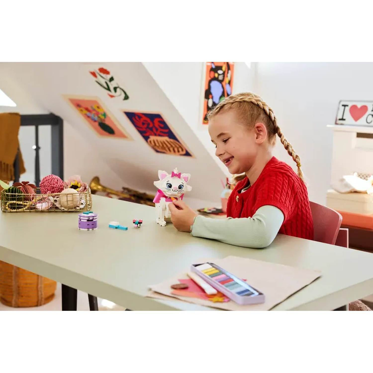 Een meisje met vlechten houdt een wit kattenspeeltje vast. Een tafel heeft kunstbenodigdheden en ander speelgoed. Kunstwerken hangen aan de muur.