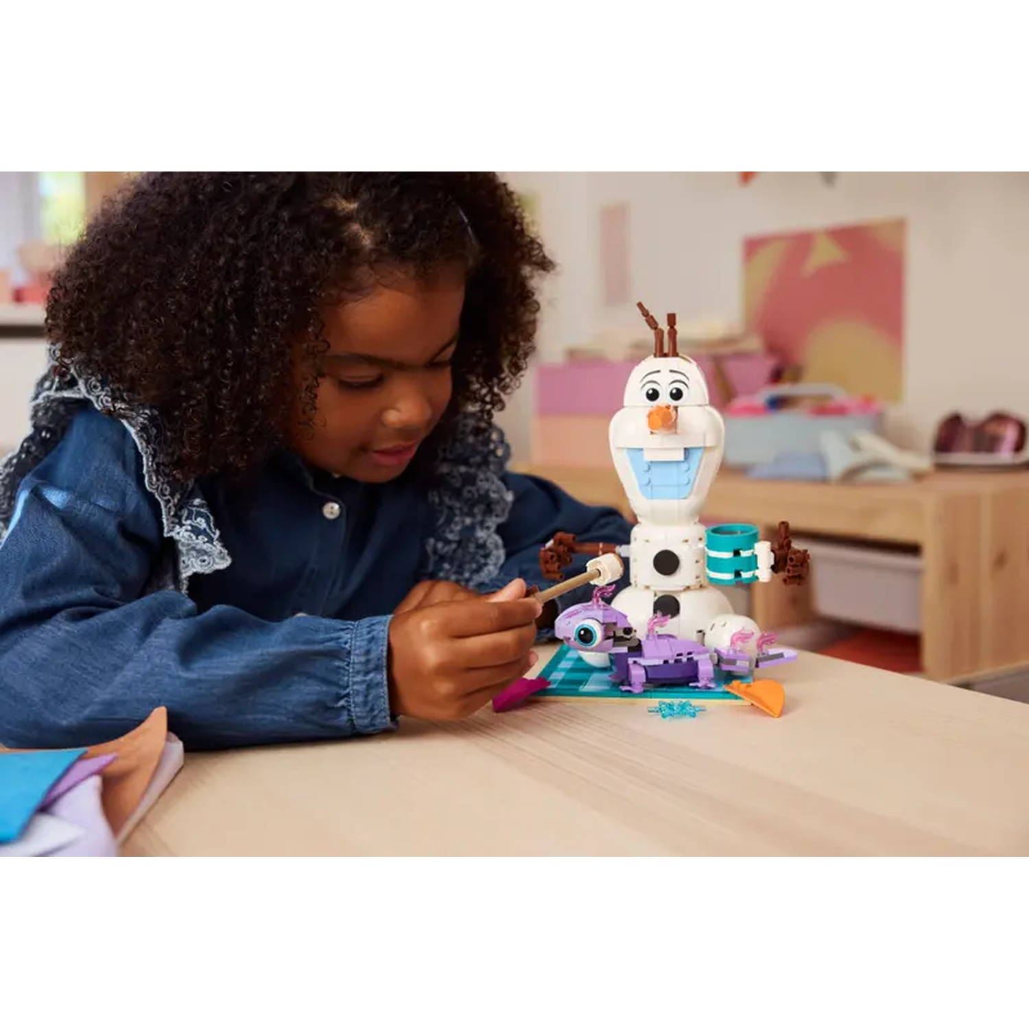 Een meisje bouwt een Lego Olaf. Ze is geconcentreerd, houdt een stok vast, naast de sneeuwman. De achtergrond omvat een tafel en een roze kunstwerk.